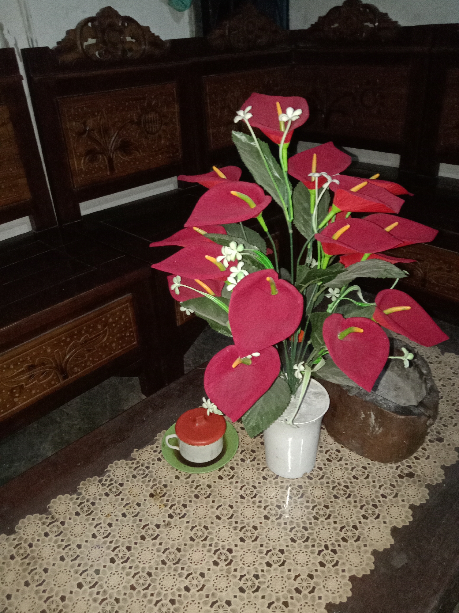 The image captures a still life scene, likely indoors, featuring a bouquet of artificial red anthurium flowers in a white pot placed on a dark wooden table. The table is adorned with a cream-colored, intricately patterned lace doily. To the left of the floral arrangement, a small white teacup with a red lid sits on a green saucer. The background shows dark wooden furniture with carved details, suggesting a traditional or antique setting. The lighting is dim, casting shadows and highlighting the textures of the objects. No people are visible in the image. The location is described as being in the city of Jumput, Indonesia, although the image itself does not contain any specific visual cues to confirm this location.