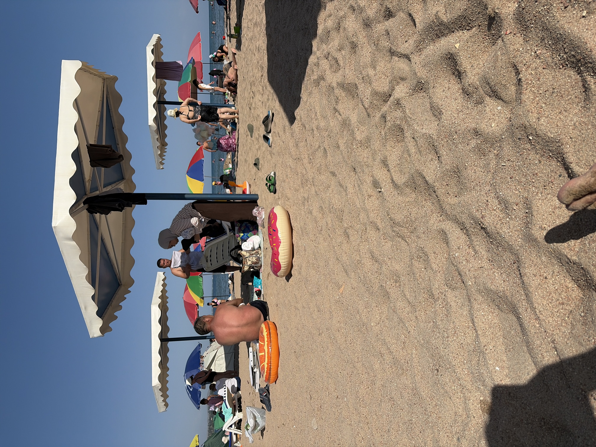 The image depicts a sunny day at a crowded beach in Cholpon-Ata, Kyrgyzstan, situated along the shores of Lake Issyk-Kul. Numerous beach umbrellas of various colors, predominantly white, blue, red, and yellow, provide shade for beachgoers. People are scattered across the sandy terrain, some sitting on lounge chairs, others standing, and a few are lying down.
In the foreground, two inflatable rings are placed on the sand, one orange and shaped like a life preserver, and the other pink and shaped like a donut. Several pairs of flip-flops are also visible on the sand, including a green pair and a blue and black pair.
A man with his back to the camera is seated on the sand, wearing dark shorts. Nearby, a woman wearing a hijab and striped top is interacting with another person holding a piece of paper. Further back, more individuals are gathered under umbrellas, with some in the water. The overall scene is characteristic of a popular tourist destination during peak season, with a vibrant atmosphere and clear blue skies. The presence of beach accessories like inflatables and umbrellas suggests recreational activities associated with a lakeside beach.