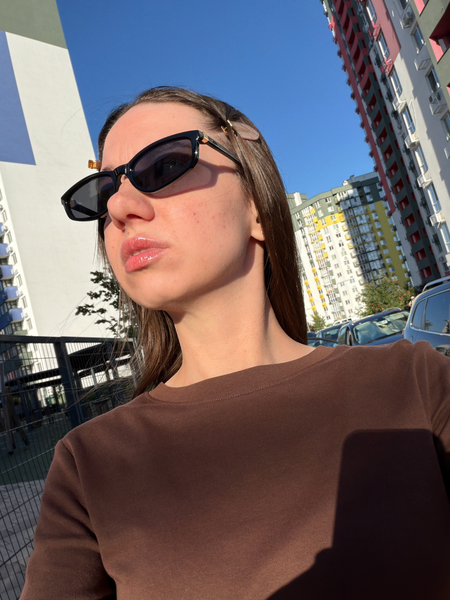 A young woman with long, dark brown hair is standing outdoors in Kyiv, Ukraine, under a clear blue sky. She is wearing black sunglasses and a brown long-sleeved shirt. Her lips are glossy and slightly parted. In the background, modern residential buildings with colorful facades and balconies are visible. Cars are parked in the foreground, and a fence surrounds a playground area. The image is taken from a low angle, looking up at the woman and the buildings.