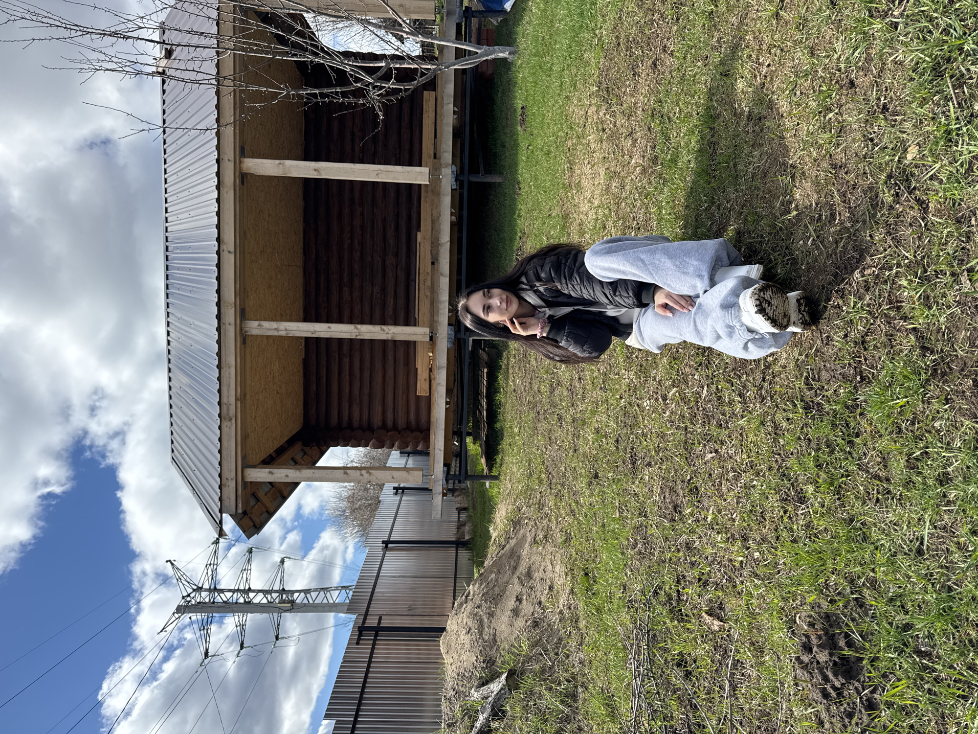 A young woman lies on the grass in Chaikovsky, Russia, in an outdoor setting. She is wearing a black jacket and gray sweatpants, and her shoes are dirty. A partially constructed log cabin with a corrugated metal roof stands to her left. Behind the cabin is a power line. A metal fence is adjacent to the cabin, with exposed dirt between the fence and the grass. The sky is visible with scattered clouds. The grass appears to be a mixture of green and brown, indicating early spring.