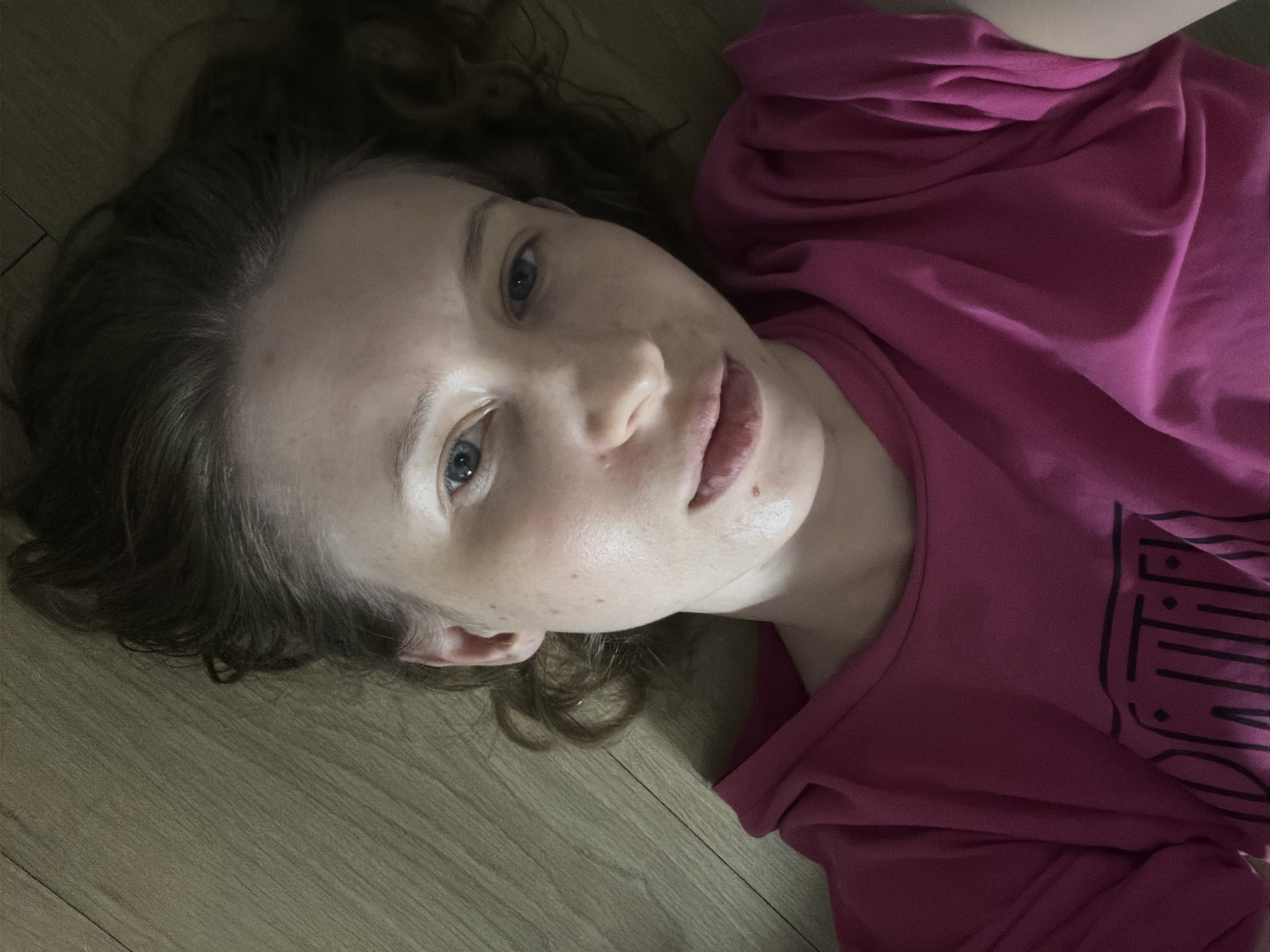 A person is lying on a wooden floor, wearing a bright pink t-shirt. The t-shirt has a black graphic design on the front. The person's face is partially visible, and they have light-colored hair. The background is a plain wooden floor. The image does not contain information about the city of Dongducheon-si, South Korea.