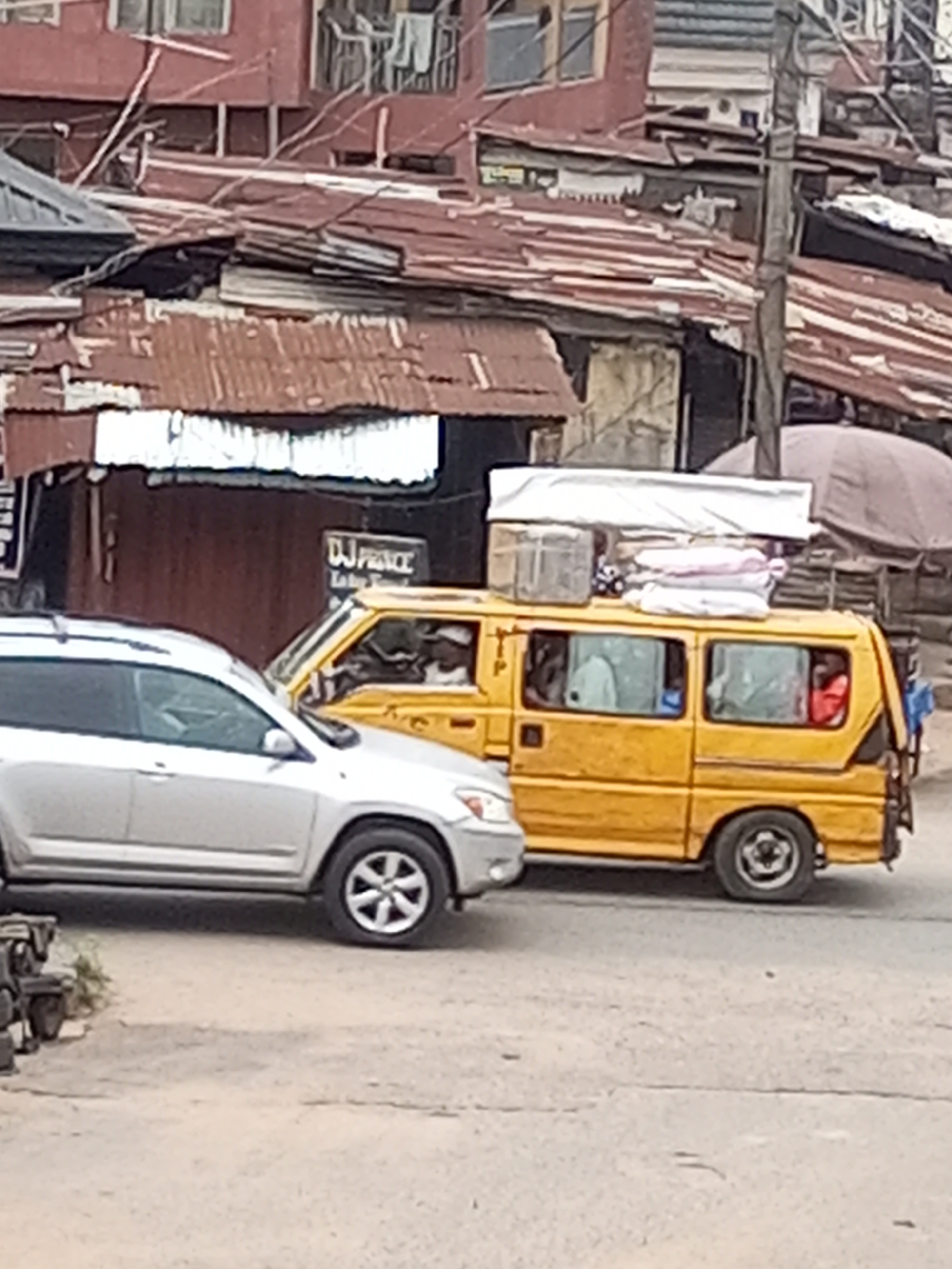 The image depicts a street scene in Onitsha, Nigeria. A silver SUV is parked on the left side of the frame. To the right, a yellow commercial minibus is visible, with luggage and other items strapped to its roof. Passengers are visible inside the bus. The background shows densely packed buildings with corrugated iron roofs. A sign is partially visible, reading "DJ Prince." The street appears to be unpaved.