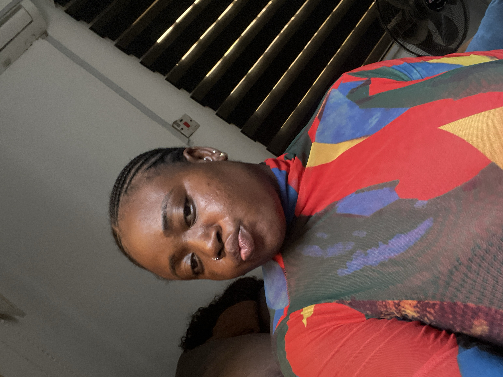 A medium shot captures a dark-skinned individual with braided hair and a nose piercing. The person is wearing a multi-colored top, primarily red, blue, green, and yellow. The individual is indoors, as evidenced by the ceiling vents and a fan visible in the background. The scene is likely in Abuja, Nigeria, as requested. Other elements in the environment include a wall with an electrical outlet.