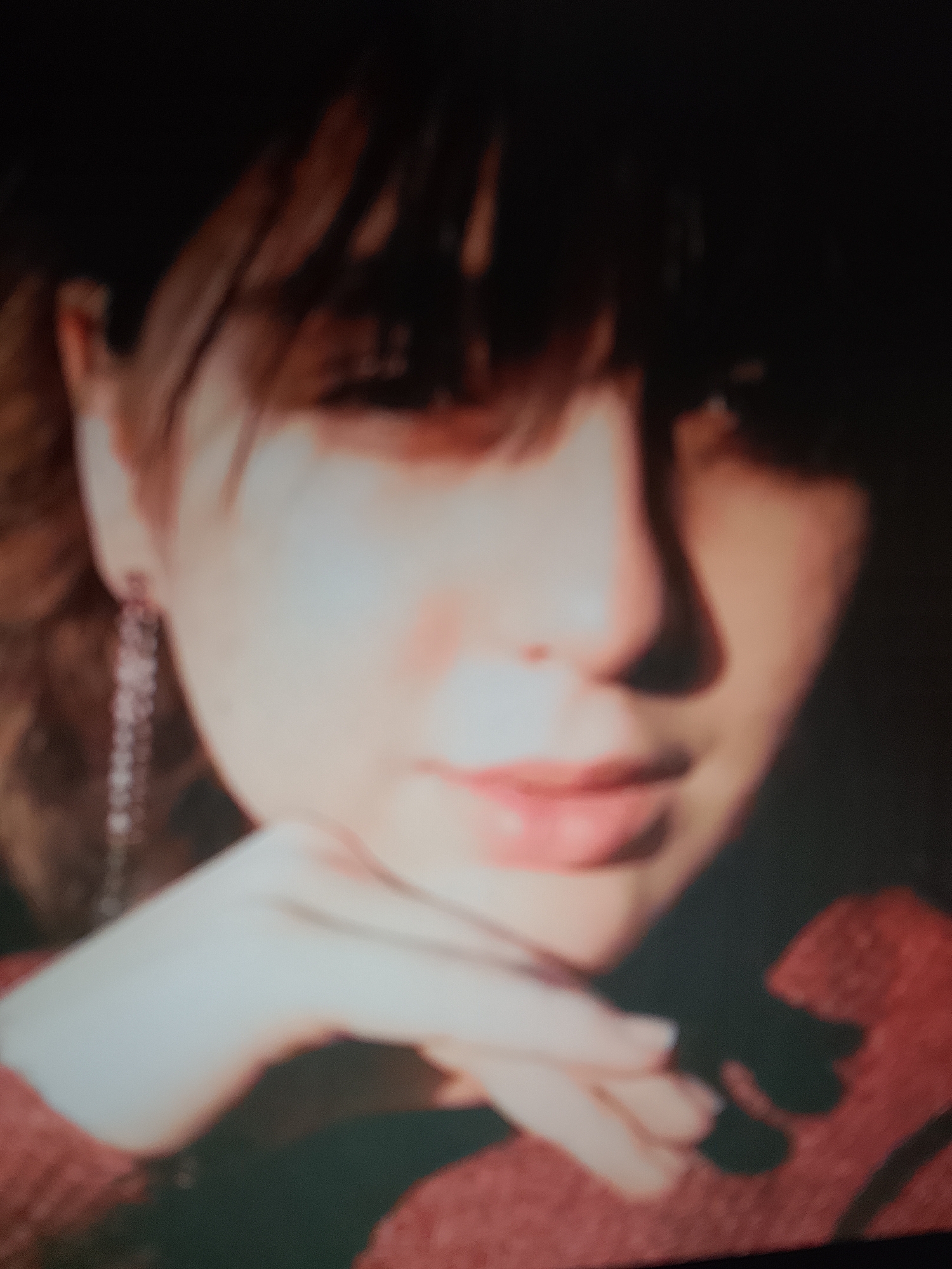The image is a close-up, out-of-focus portrait of a young woman. She has light-colored skin, brown hair with bangs, and is wearing a red garment. Her left hand is resting on her chin. A long, dangling earring with multiple strands is visible on her left ear. The background is dark and indistinct. There is no discernible information about the location in Ostróda, Poland.