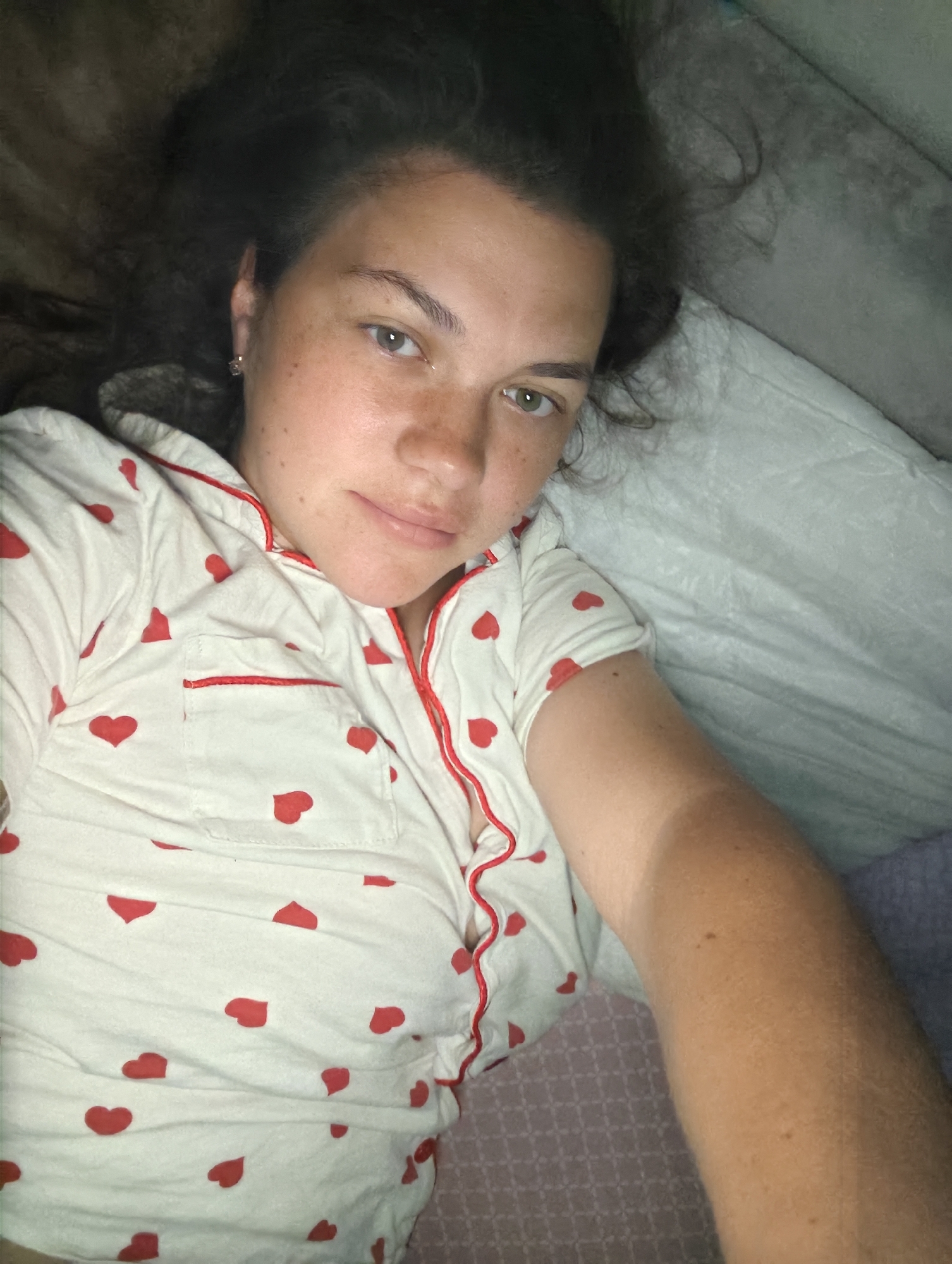 A young adult female is depicted lying in bed, looking directly at the camera, likely in a selfie or close-up shot from above. She has dark, curly hair and light-colored eyes, appearing to be greenish-grey. Her face and arms display numerous freckles.
She is wearing white pajamas patterned with small red hearts, featuring red piping along the collar and front placket. A small chest pocket, also outlined in red, is visible on her left side. A small, clear or diamond-like earring adorns her left earlobe. Her expression is neutral, with a soft, closed-mouth smile.
The setting is an indoor bedroom, with various bedding visible. A white pillow supports her head and upper body. Behind her head and to her left, there's a dark, textured blanket or headboard. A light grey sheet or blanket covers a portion of her right arm, which shows a distinct tan line. A patterned pinkish-purple sheet or blanket is visible beneath her left arm. The lighting in the scene is dim, suggesting it could be nighttime or a softly lit room. There is no visible text in the image.