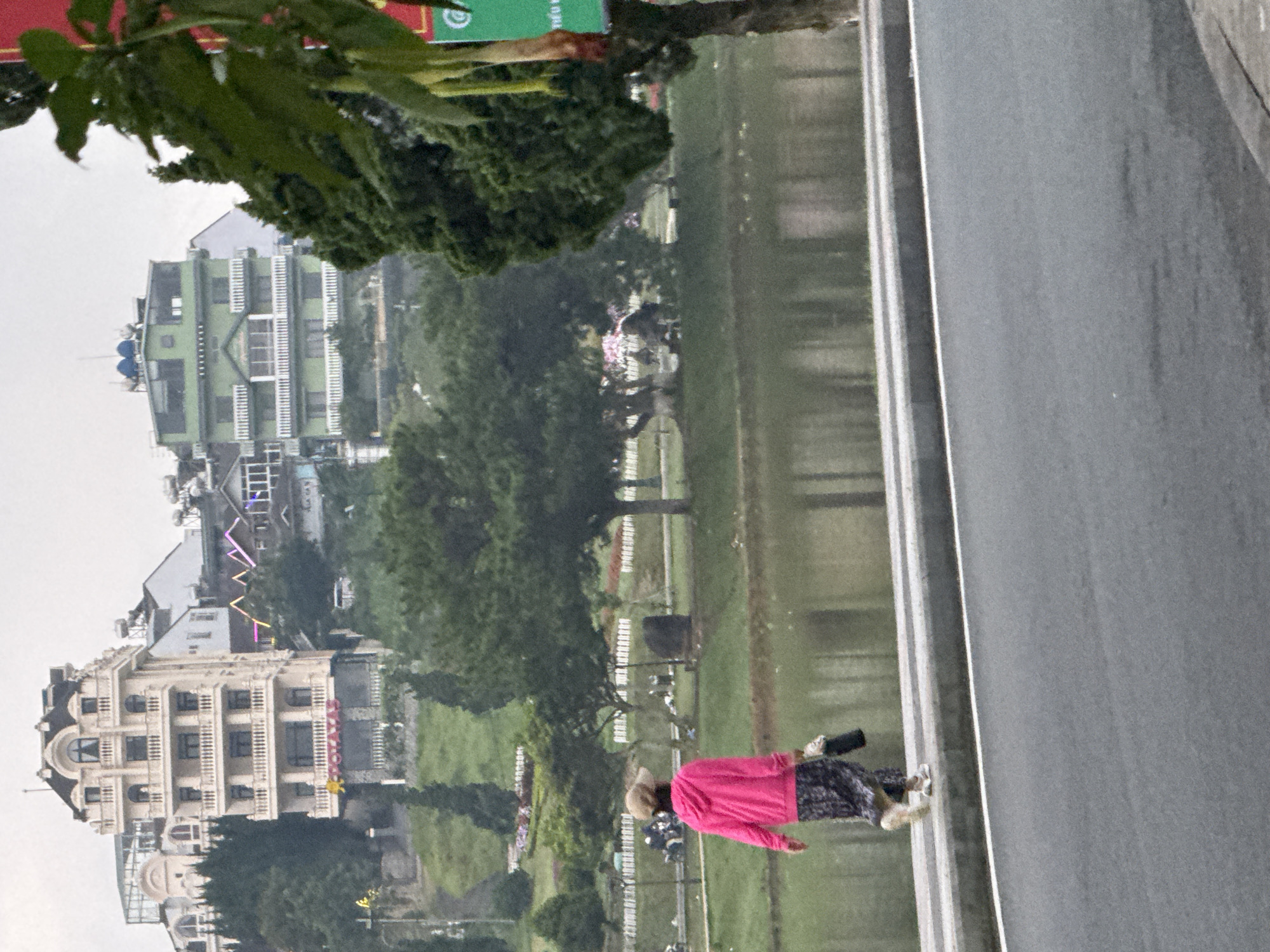 The image depicts a cloudy day scene in Xuân Hương Lake, Đà Lạt, Vietnam. A person wearing a pink hooded jacket and a patterned skirt walks along a paved pathway next to the lake. They are carrying a dark bag in their right hand. In the background, several multi-story buildings are visible on a hillside, with trees interspersed between them. One building on the left is ornate and cream-colored, while a building in the middle is painted bright green. The lake's surface is calm, reflecting the overcast sky. The foreground shows a grey asphalt road with a white line marking the edge of the pedestrian pathway.