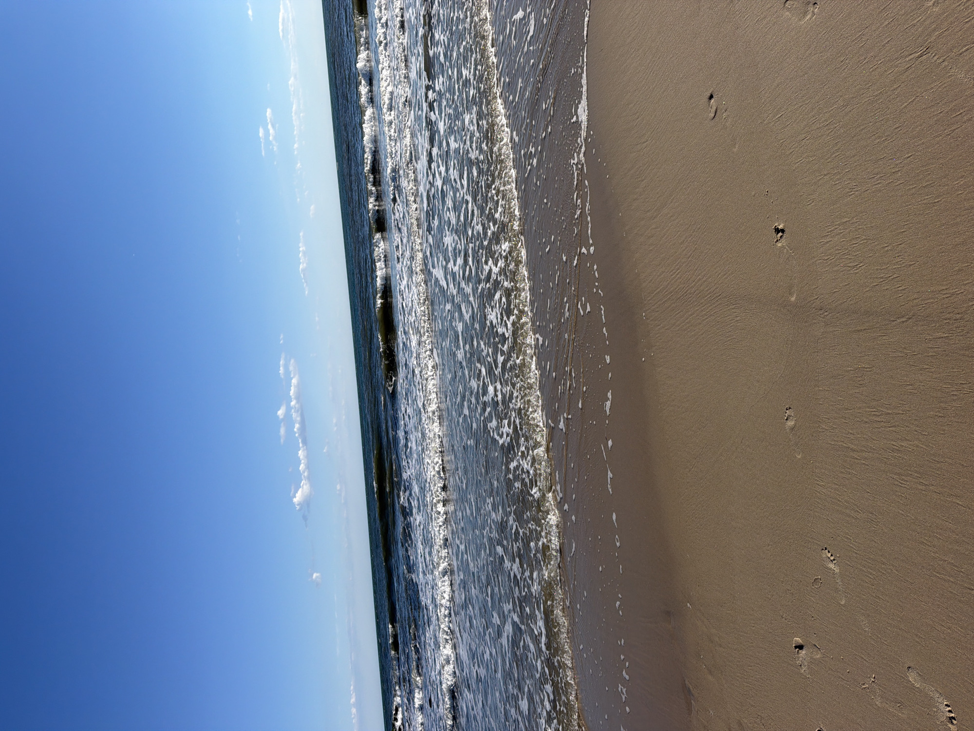 The image depicts a serene beach scene in Hampton Bays, United States of America. The foreground is dominated by a sandy beach with visible footprints, suggesting recent human presence. Gentle waves lap at the shore, leaving a trail of foam and glistening water on the wet sand. The ocean stretches out to the horizon, its surface rippling under a clear blue sky dotted with a few wispy white clouds. The overall impression is one of a tranquil and picturesque coastal environment.