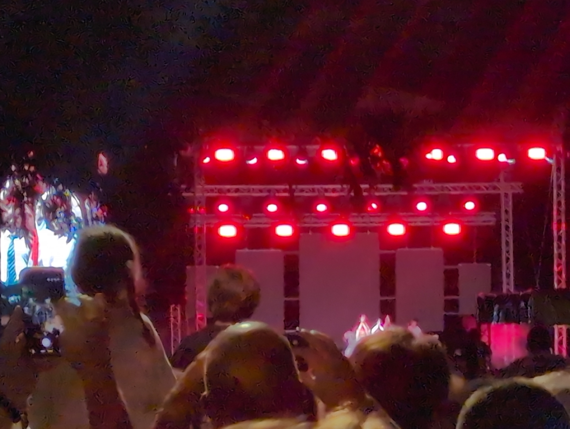 The image shows a crowd of people at an outdoor event at night, illuminated by bright red stage lights. The foreground is filled with the backs of people's heads and shoulders, some holding up mobile phones to capture the event. One person in the mid-left is holding a phone with a visible screen. In the background, a stage structure is visible with multiple rows of bright red lights. Two large, pale rectangular screens or backdrops are positioned on the stage. Several figures can be seen on the stage, appearing to be performers, though they are indistinct due to distance and lighting. The event appears to be a concert or performance. The lighting suggests it is taking place in Varna, Bulgaria, likely during a summer festival or open-air concert.