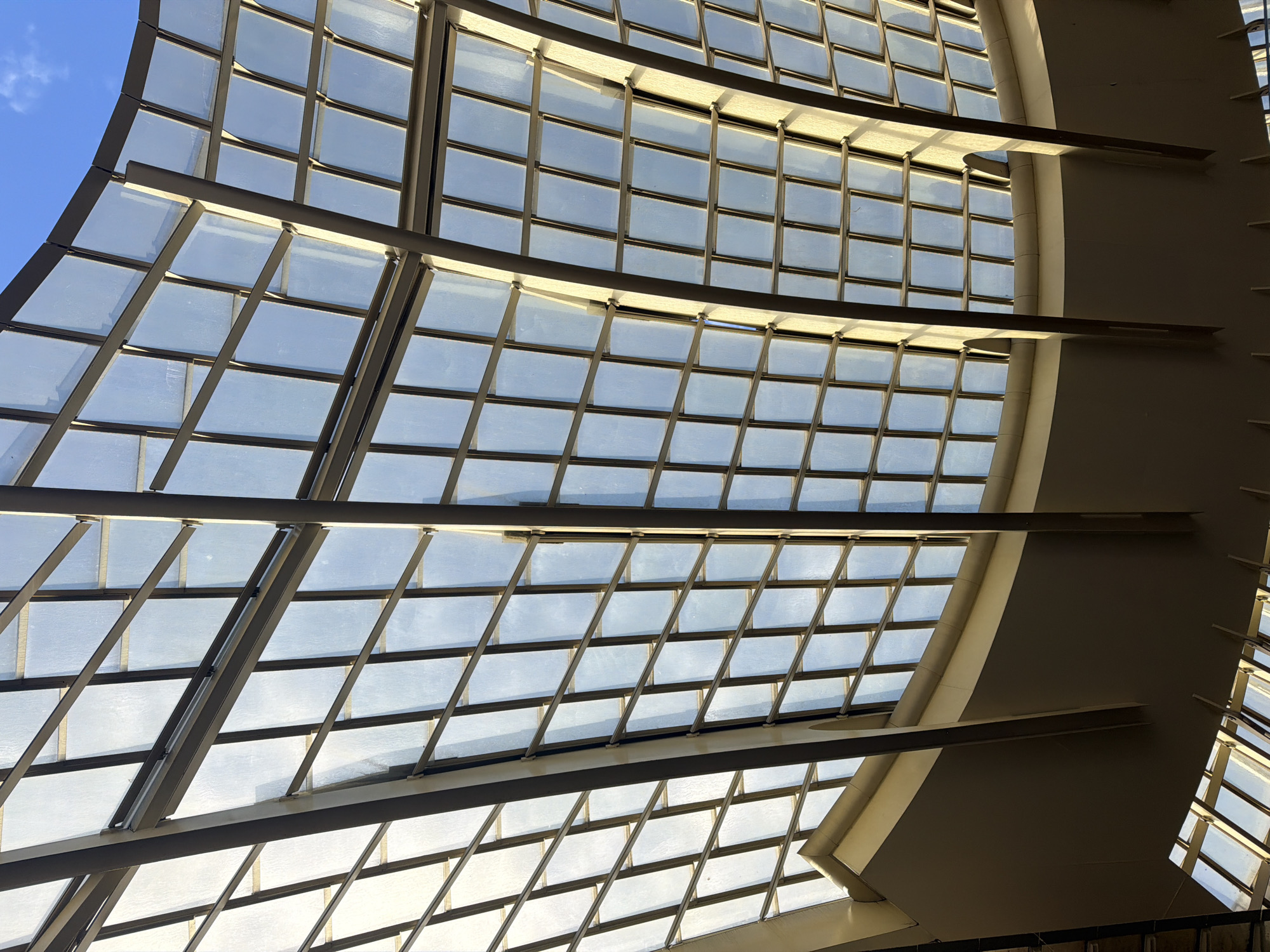 The image displays an upward view of a large, curved architectural ceiling structure within the Grande Arche de la Défense, located in Puteaux, a suburb immediately west of Paris, France, forming part of the La Défense business district. The ceiling is primarily composed of a grid of numerous rectangular glass panels, each framed by a dark metal skeleton, likely steel or aluminum. Through the glass, a clear blue sky with sparse, faint white clouds is visible, indicating an exterior view above the structure. Natural light illuminates the structure, creating distinct patterns of light and shadow on the metal framework and the internal surfaces. Towards the bottom and mid-sections of the frame, parts of a solid, light-beige internal architectural element, possibly a walkway or support structure, are visible with a curved underside and vertical beams extending into the glass canopy. No individuals or discernible actions are present within the captured scene.