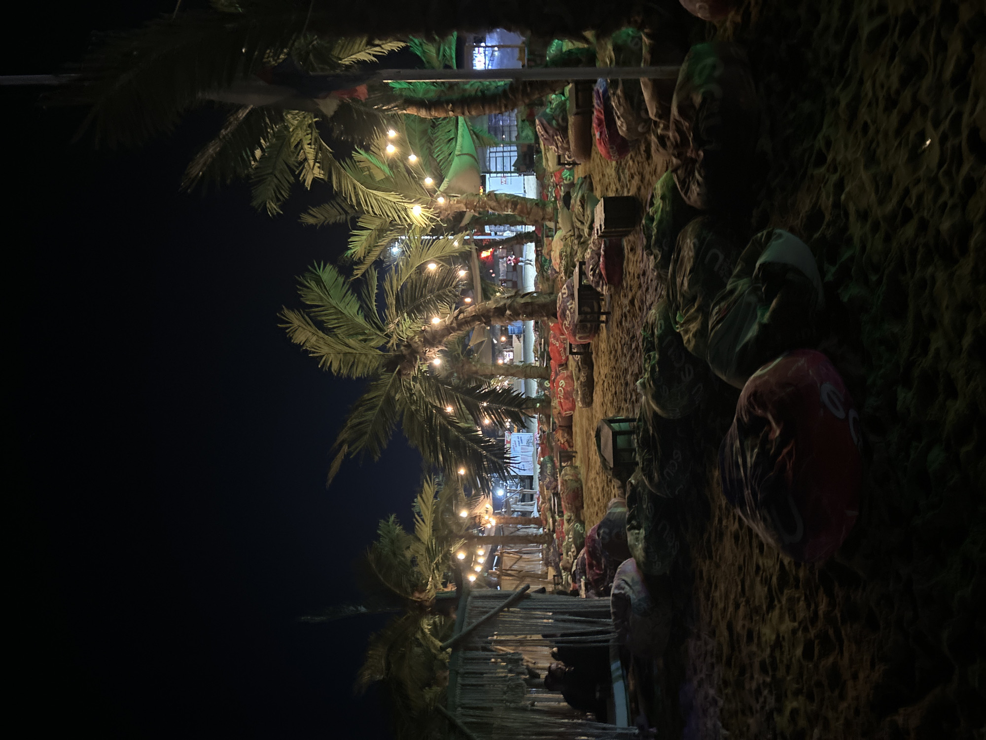 The image shows an outdoor scene at night, potentially on a beach in the city of Nesebar, Bulgaria. A row of palm trees, adorned with string lights, lines the left side of the image, leading into the distance. The trees cast shadows on the sandy ground. To the right of the trees, a row of colorful bean bag chairs and small tables suggests an outdoor seating area for a bar or restaurant. The background features a brightly lit structure. The lighting is dim and atmospheric.