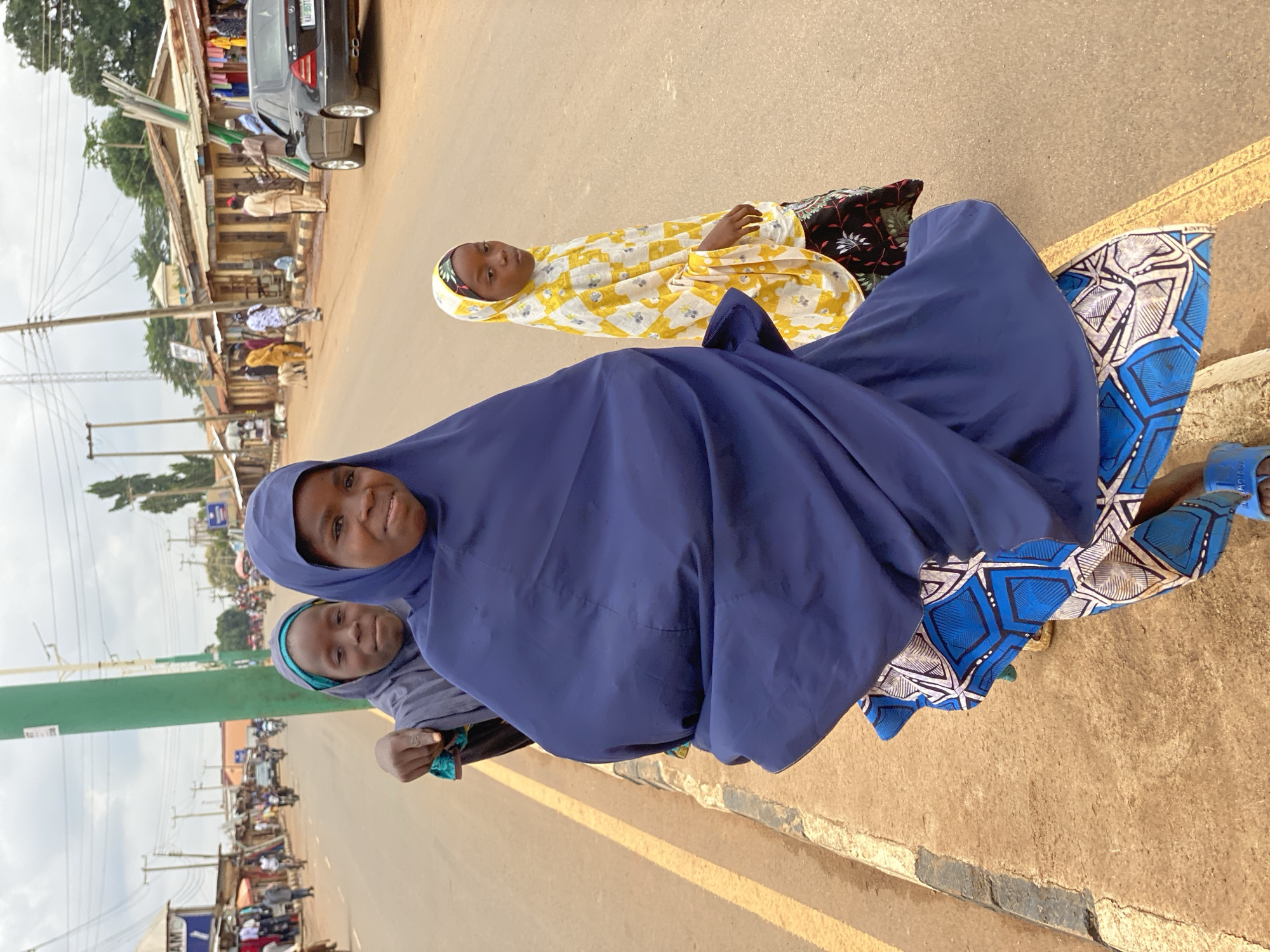 Three females are on a street. Two females are wearing hijabs. One female is wearing a yellow and white top with black pants. The females are on a street in Kafanchan B, Nigeria. There is a black car parked along the street. There are buildings in the background.
