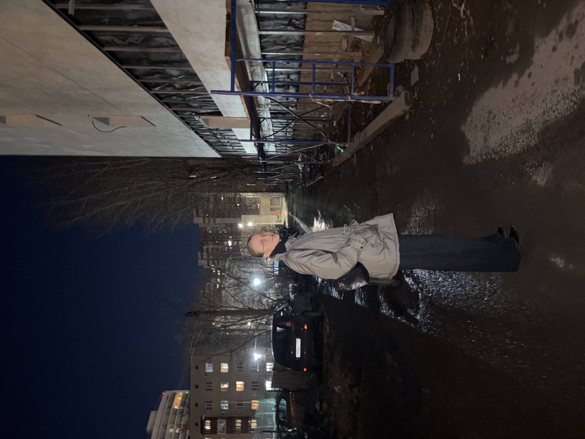 The image, taken at night, depicts a street scene in what is likely Perm, Russia, based on the car license plate. A person stands on the wet pavement, wearing a light grey jacket and dark pants. Behind the person is a dark-colored car, with the license plate number "CH150EY192" visible. In the background, tall apartment buildings are illuminated with lights. Bare trees are visible. A building under construction is on the left of the frame with scaffolding and construction materials.