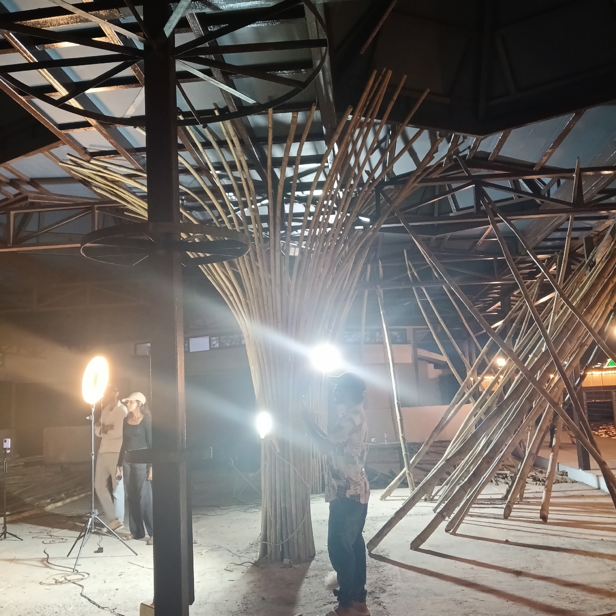 The photo shows an interior shot of a structure under construction. It is located in Panaji, India, based on metadata. A metal framework forms the roof, with bamboo poles arranged artistically as a central pillar and additional supporting structures. Three people are present: one person is standing next to the central pillar, perhaps inspecting it, while two others stand near a light source mounted on a tripod. The ground appears to be concrete, and several objects, including construction materials and equipment, are scattered throughout the space.