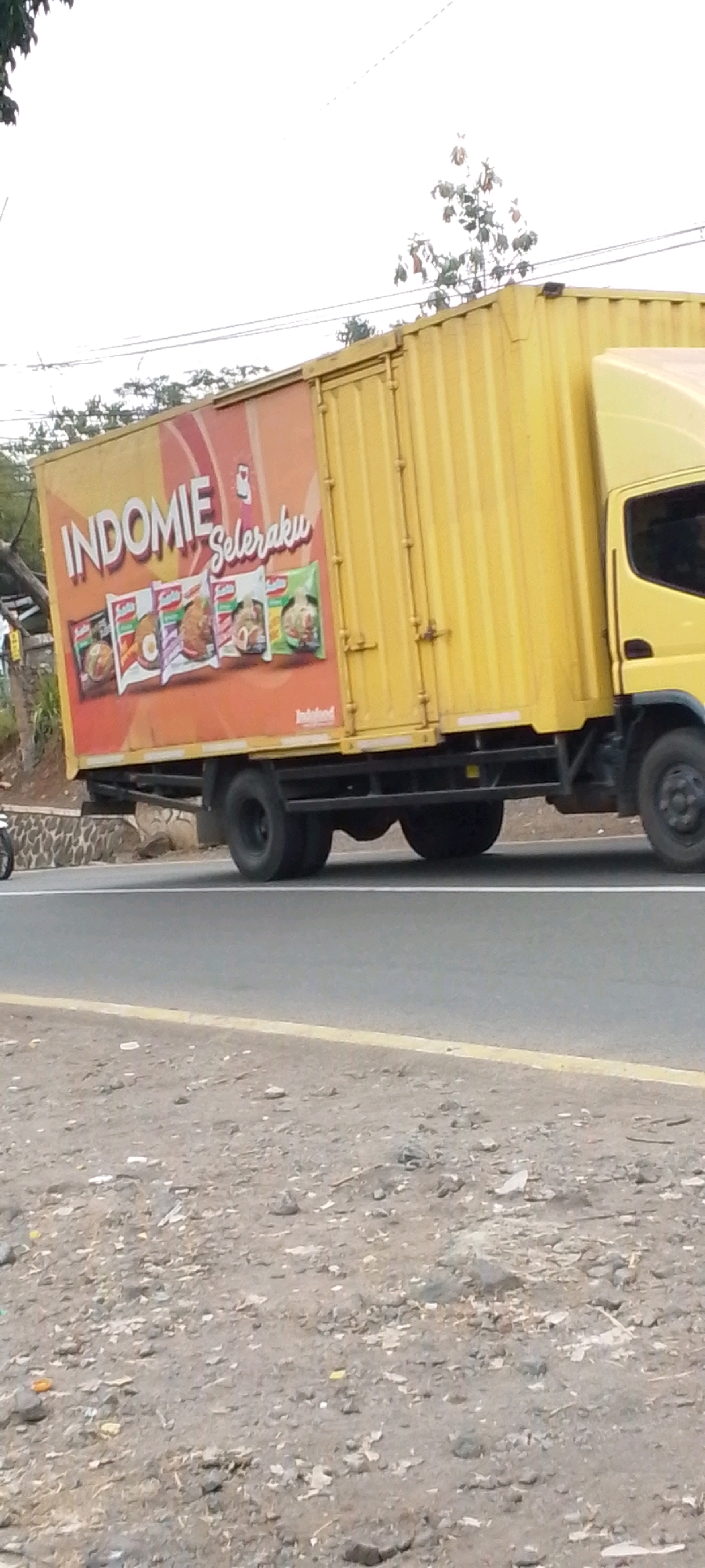A yellow box truck is parked on the side of a road in Cilaku, Indonesia. The truck is branded with "INDOMIE SeleraKu" and features images of various Indomie noodle products. The truck is a commercial vehicle, likely used for transporting goods. The road appears to be paved and there is gravel and debris on the shoulder. Trees and foliage are visible in the background. The sky is overcast.