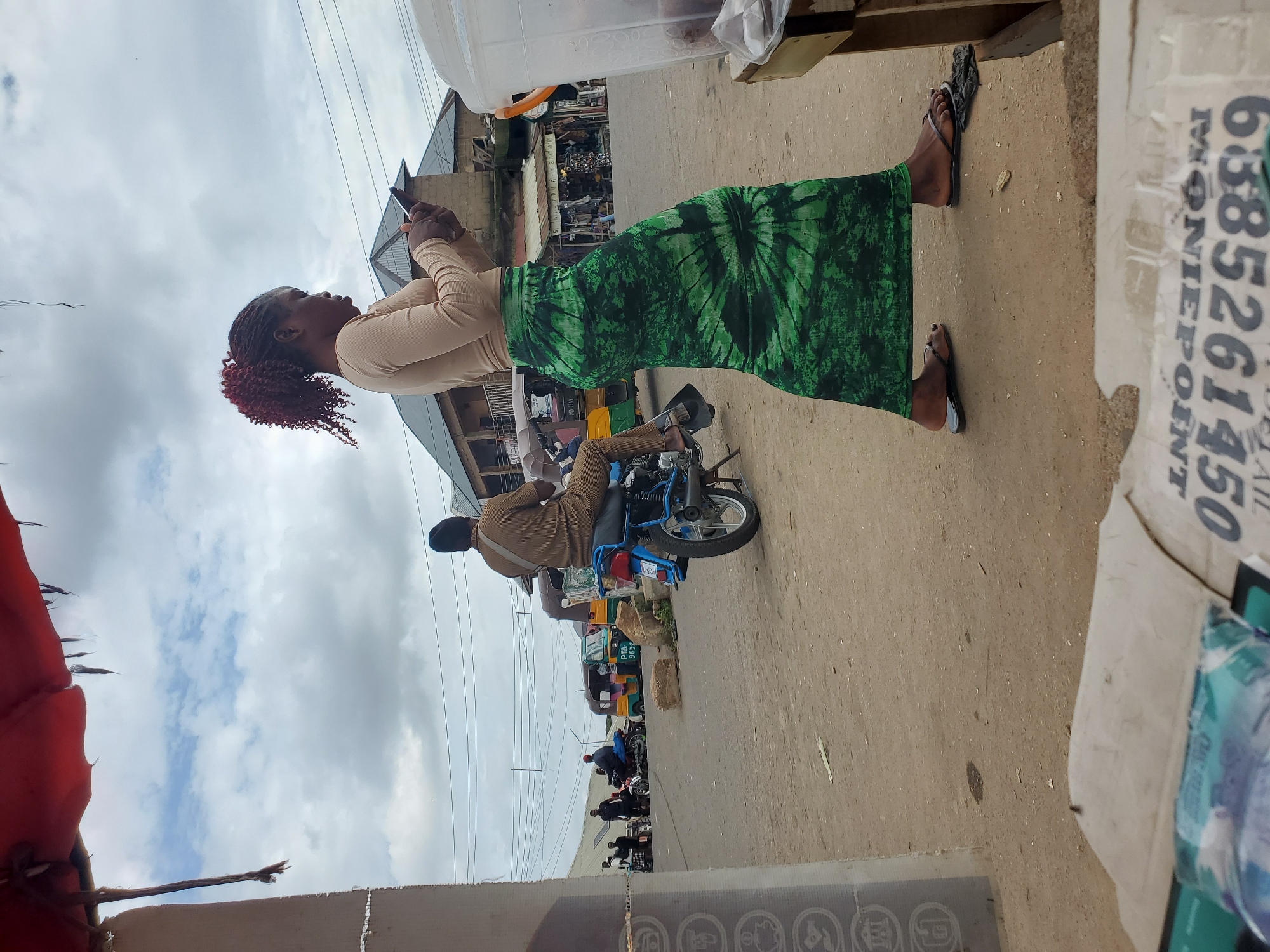 In Gwong, Nigeria, under a cloudy sky, a woman with braided auburn hair, wearing a beige long-sleeved shirt and a green tie-dye maxi skirt, stands facing to her left, engrossed in her smartphone. Beside her, a large clear plastic container sits on a wooden stall. Behind her, a man on a blue motorcycle, dressed in a striped brown shirt and dark pants with a blue cap, is looking away from the camera. In the background, several auto-rickshaws and motorcycles are visible, along with buildings and power lines. The ground is dusty and uneven, with scattered debris. In the foreground, partially visible, are pieces of cardboard and packaging.