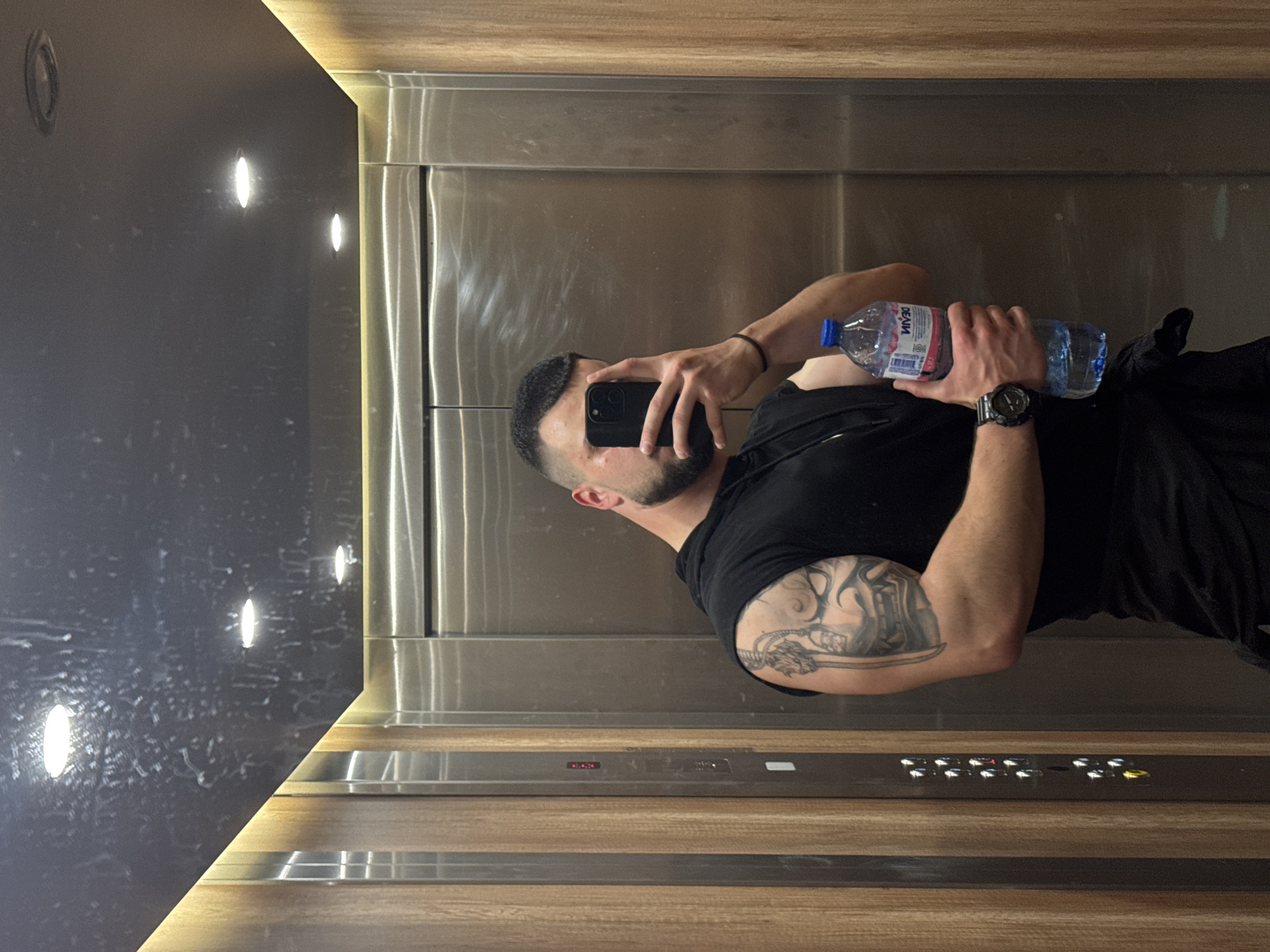 The image, originally oriented horizontally, depicts a man taking a mirror selfie inside an elevator. The man is positioned towards the center-right of the frame, seen in reflection. He has a dark beard and short hair, and is wearing a black t-shirt. A prominent tattoo featuring a design that includes a sword or similar element is visible on his left upper arm. He wears a dark wristwatch on his left wrist and a black wristband on his right.
In his right hand, he holds a dark-colored smartphone, aimed to capture his reflection. His left hand holds a clear plastic water bottle with a blue cap. The bottle's label clearly displays the text "ДЕВИН" (DEVIN), "Оригинална" (Original), and "Вода" (Water), along with "1.5L".
The elevator interior features brushed stainless steel doors and a reflective surface, which serves as the "mirror" for the selfie. To the left, a dark grey or black wall is visible, equipped with several recessed, oval-shaped lights emitting bright white light. The top and bottom sections of the elevator's interior appear to be paneled with light brown wood, featuring horizontal metallic accents on the lower panel. On the lower right, an elevator control panel is visible, displaying the number "3" in red digital text, likely indicating the current floor, along with an array of illuminated buttons. The overall setting appears modern and clean. Given the visible text on the water bottle, the location is consistent with Sofia, Bulgaria.