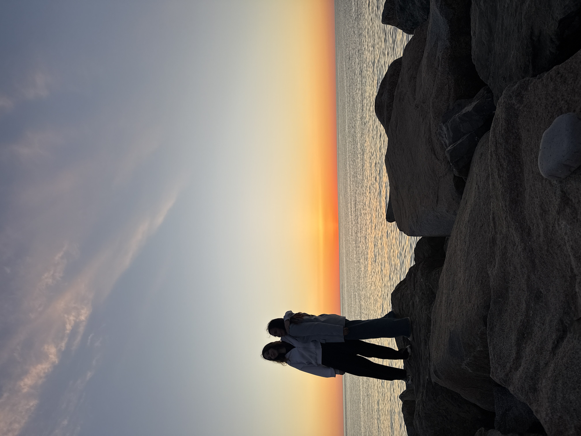 Two figures stand on a cluster of dark, large rocks at the edge of a body of water, likely the Black Sea. The figures appear to be young women, seen in silhouette, wearing light-colored hoodies and dark pants. They seem to be standing close to one another, possibly embracing. The background displays a vivid sunset with a gradient of colors, from a pale yellow near the horizon to orange and then a soft blue higher in the sky. This scene is likely set in Batumi, Georgia, a city known for its coastal location along the Black Sea and its dramatic sunsets. The rocks appear to be part of a man-made breakwater or jetty. The water reflects the colors of the sunset.