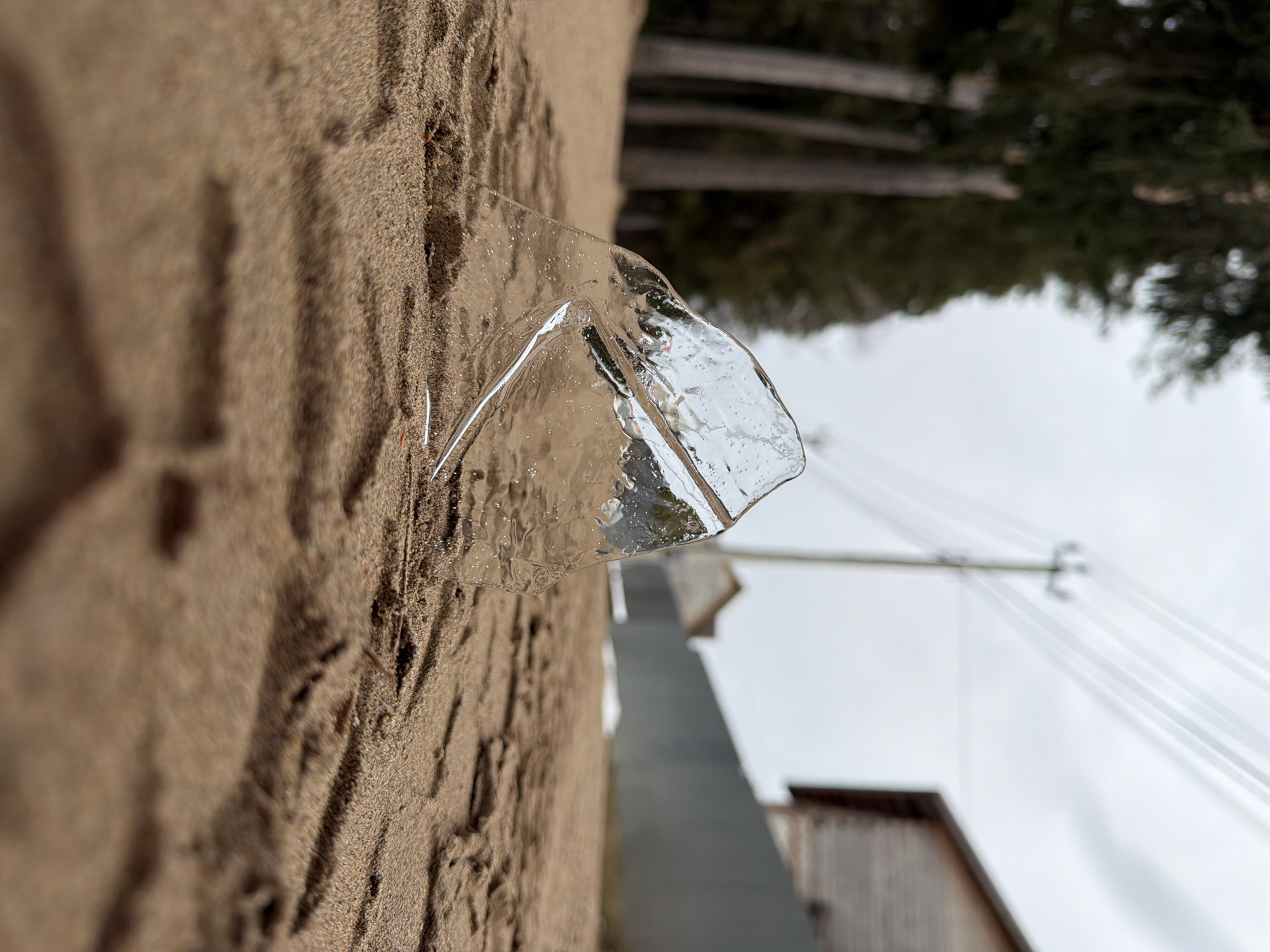 The image shows a close-up of a piece of ice resting on a sandy surface. The background includes trees, utility poles and lines, and parts of buildings. The location is Chaikovsky, Russia. The image appears to have been rotated counterclockwise, placing the horizon on the left. The ice is angular and partially clear, with internal imperfections visible. The sand surface shows textured patterns.