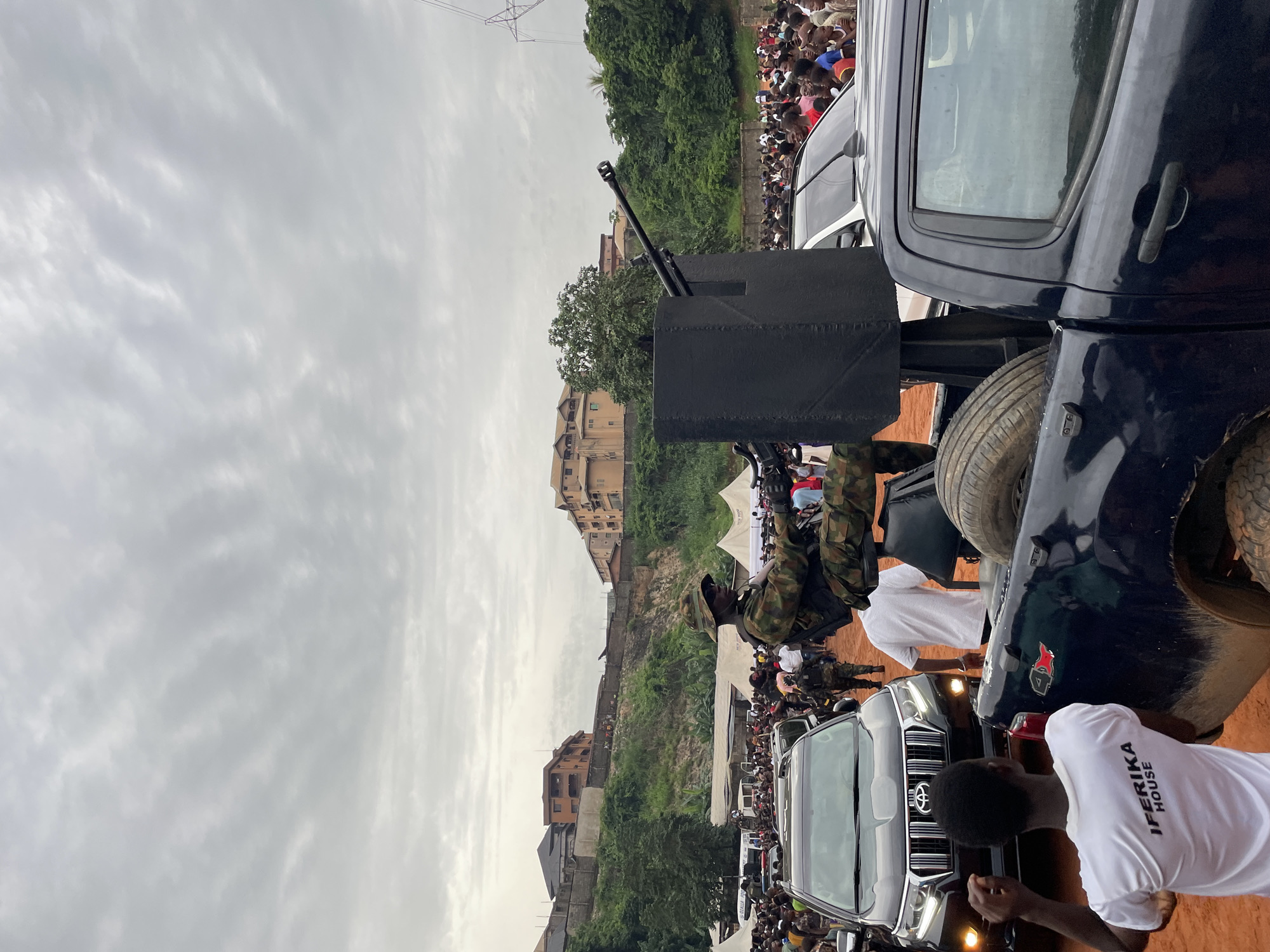 In an outdoor urban setting, a military vehicle equipped with a mounted gun is prominent in the foreground, with a soldier in camouflage uniform operating it. The vehicle is surrounded by a crowd of people, suggesting a public event or gathering. Behind the crowd, several buildings are visible on a hillside, with lush green trees interspersed among them. The sky is overcast with gray clouds. The ground appears to be reddish-brown dirt. A black SUV is parked in front of the military vehicle, and another dark-colored pickup truck is to the right. A young man in a white t-shirt with "IFÉRIKA HOUSE" printed on the back is standing in the lower left corner, facing away from the camera.