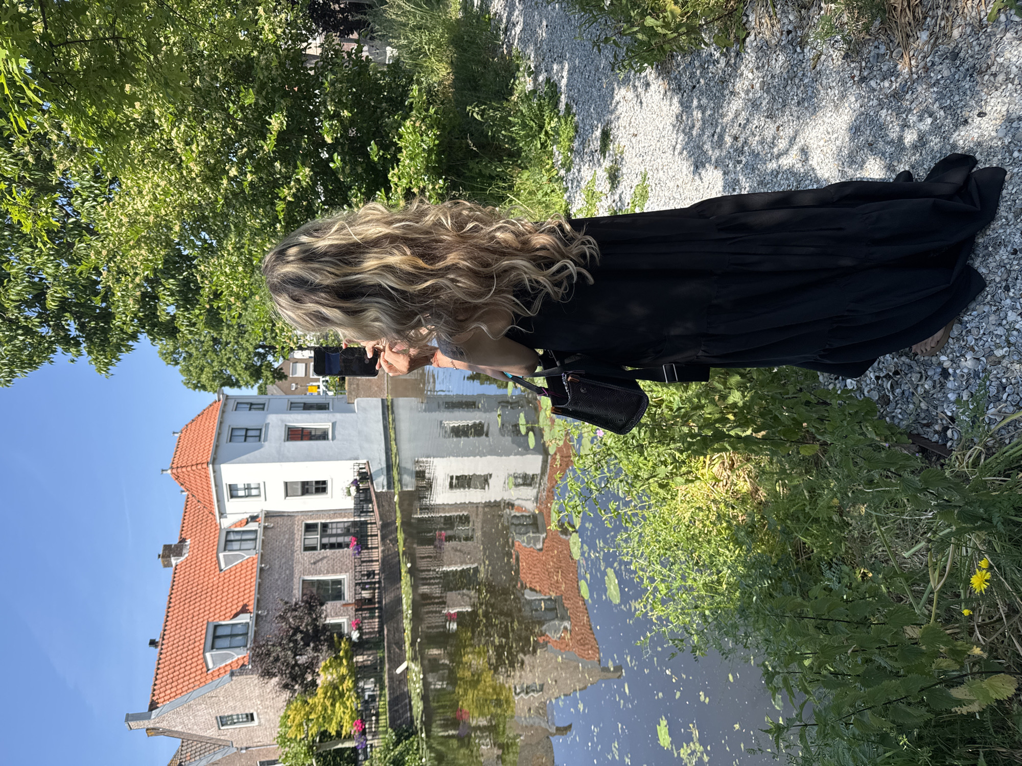 A waist-up shot shows a woman standing by a canal in Katwijk, Netherlands, taking a photo with her mobile phone. She has long, blonde, curly hair and wears a long, black, tiered dress, as well as a small, black purse over her shoulder. She stands on a path covered in gray gravel, and green vegetation is visible around her.
Across the canal, Dutch-style houses with red-tiled roofs and white walls reflect in the water. The sky is clear and blue.