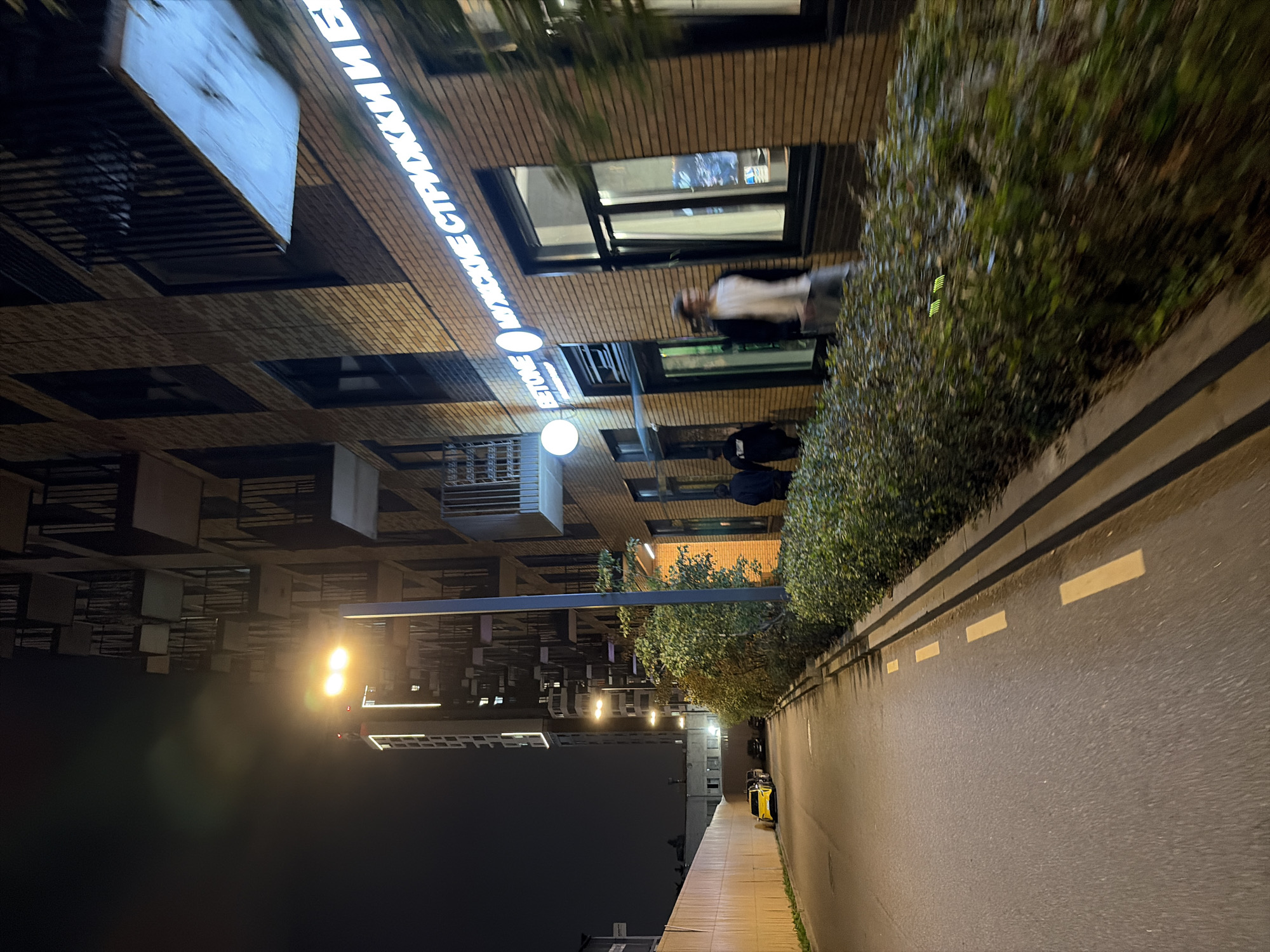 The image depicts a nighttime street scene in Moscow, Russia, with a focus on a modern building facade and an adjacent walkway. A multi-story residential building with balconies and brickwork dominates the right side of the frame, illuminated by streetlights. A neon sign, reading "BETONE МУЖСКИЕ СТРИЖКИ И БЕ," is prominently displayed on the building's exterior, indicating a men's barbershop. Several people are visible on the walkway; two figures in dark clothing are walking away from the camera, while another individual in a light-colored shirt and grey pants is walking towards the camera, slightly blurred due to motion. A street with yellow lane markings runs along the left side of the image, with a parked yellow car visible further down. The walkway is lined with manicured bushes. The overall ambiance is that of a well-lit urban environment at night.