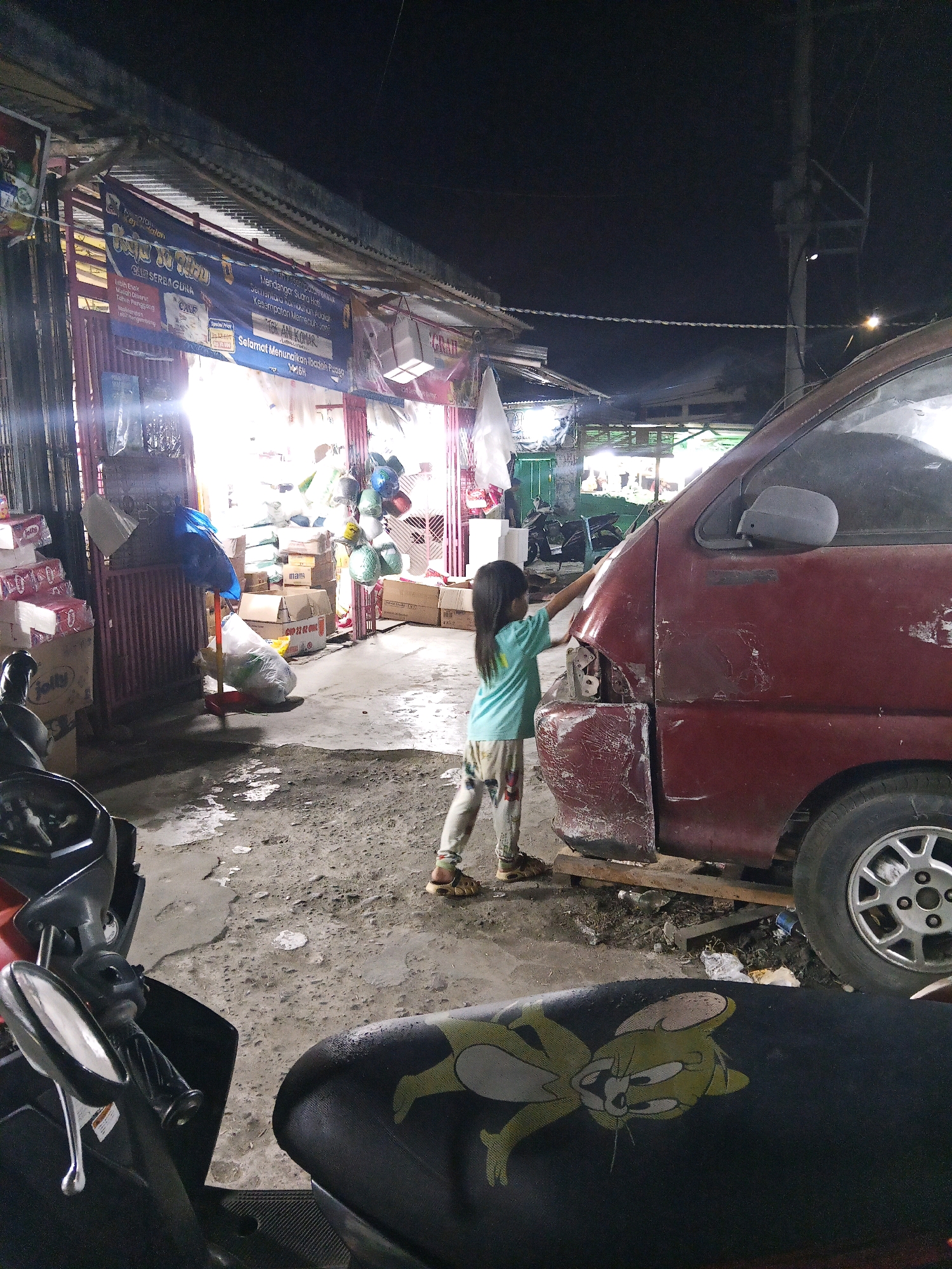 At night, a young girl with long dark hair, wearing a light blue t-shirt and patterned leggings, stands on the right side of the frame, facing a damaged maroon van. She is touching the front of the van, near the headlight area, which appears to be detached and hanging. The van is parked on a dirt and concrete surface. In the foreground, the seat of a black motorcycle is visible, with a cartoon character resembling Tom and Jerry printed on it. To the left, a stall or shop is set up, with various items, including boxes, bags, and what appear to be balloons, displayed under a corrugated metal roof. The overall environment suggests an outdoor market or roadside stall in a somewhat disorganized urban area. The banner visible above the stall has text in Indonesian. The scene is illuminated by artificial lights, indicating it is nighttime. The presence of a motorcycle and the van suggest a transportation hub or service area. The context provided points to the location being in Lubuklinggau, Indonesia, likely a commercial or residential area.