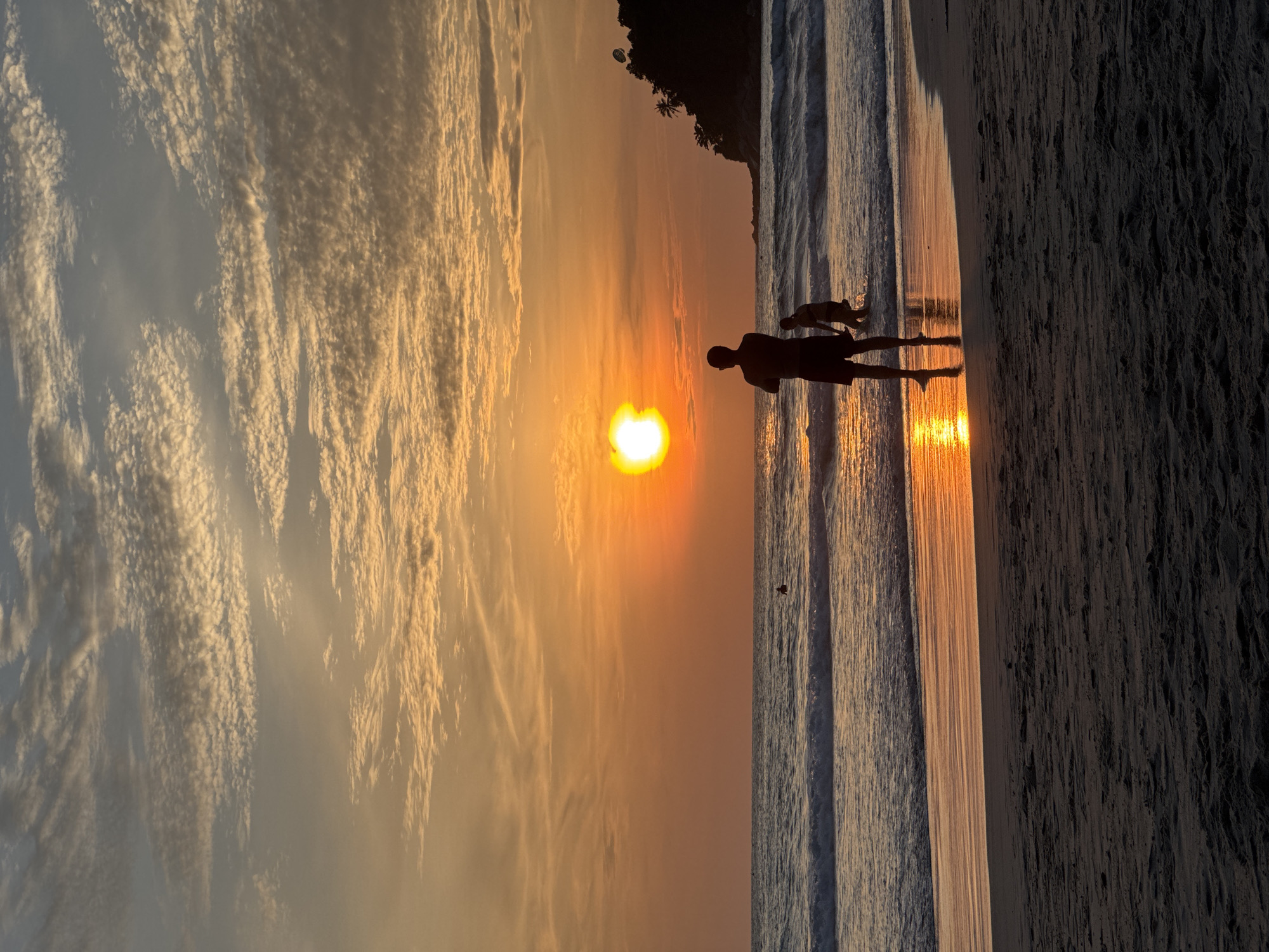 This is a vertical, eye-level, full shot of a sunset on Karon Beach, Thailand. The sun is a bright orange orb low on the horizon, casting an orange glow across the sky and reflecting on the water. Wispy, white clouds are scattered across the sky, tinged with orange and yellow from the setting sun. The ocean is calm, with small waves breaking on the shore. A silhouette of a person stands in the foreground, facing the ocean with their arms crossed. Another silhouette of a child is visible in the shallow water to the right of the person. The beach is dark sand, with the wet sand near the water reflecting the sunset colors. In the distance, a dark, tree-covered hill forms the coastline on the right.