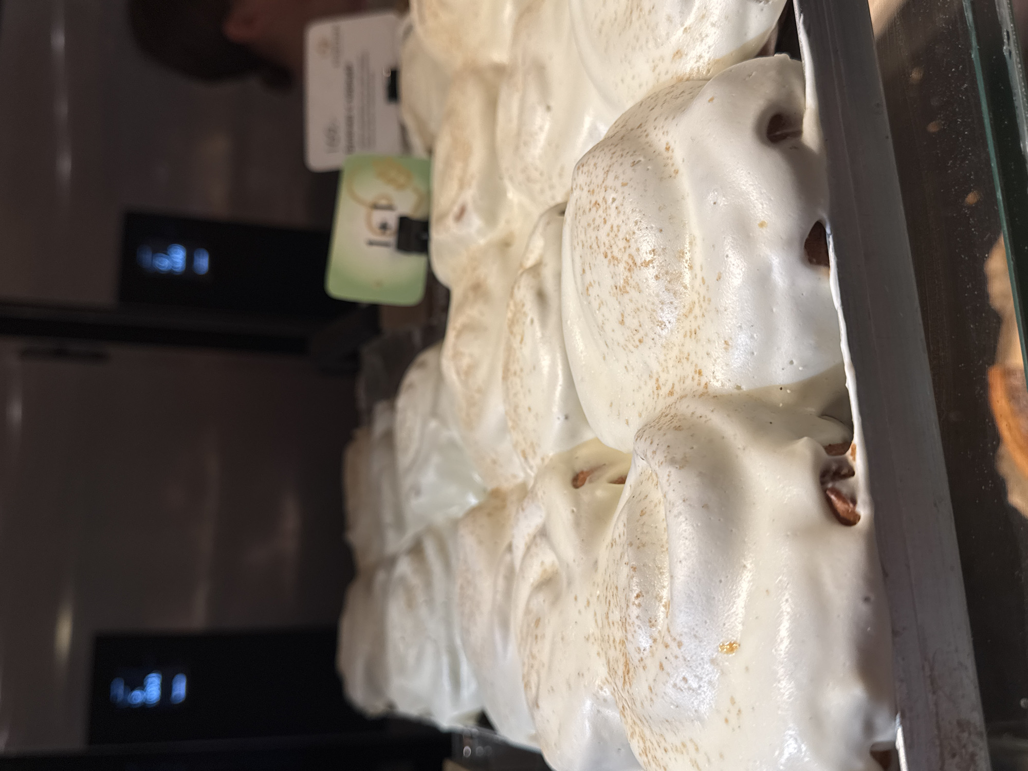 The image shows a close-up of pastries displayed in a glass-fronted case. The pastries appear to be meringue-based, with a textured white coating and speckled with brown specks. There is a price tag and a green label visible behind the pastries. The display case is situated in Izhevsk, Russia. There is a person partially visible, and a screen visible in the background.