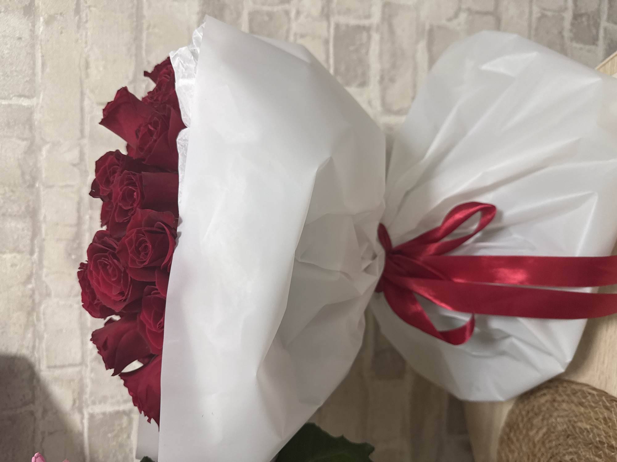 A bouquet of red roses is wrapped in white paper and tied with a red ribbon. The bouquet is placed on a wooden surface in front of a brick wall.