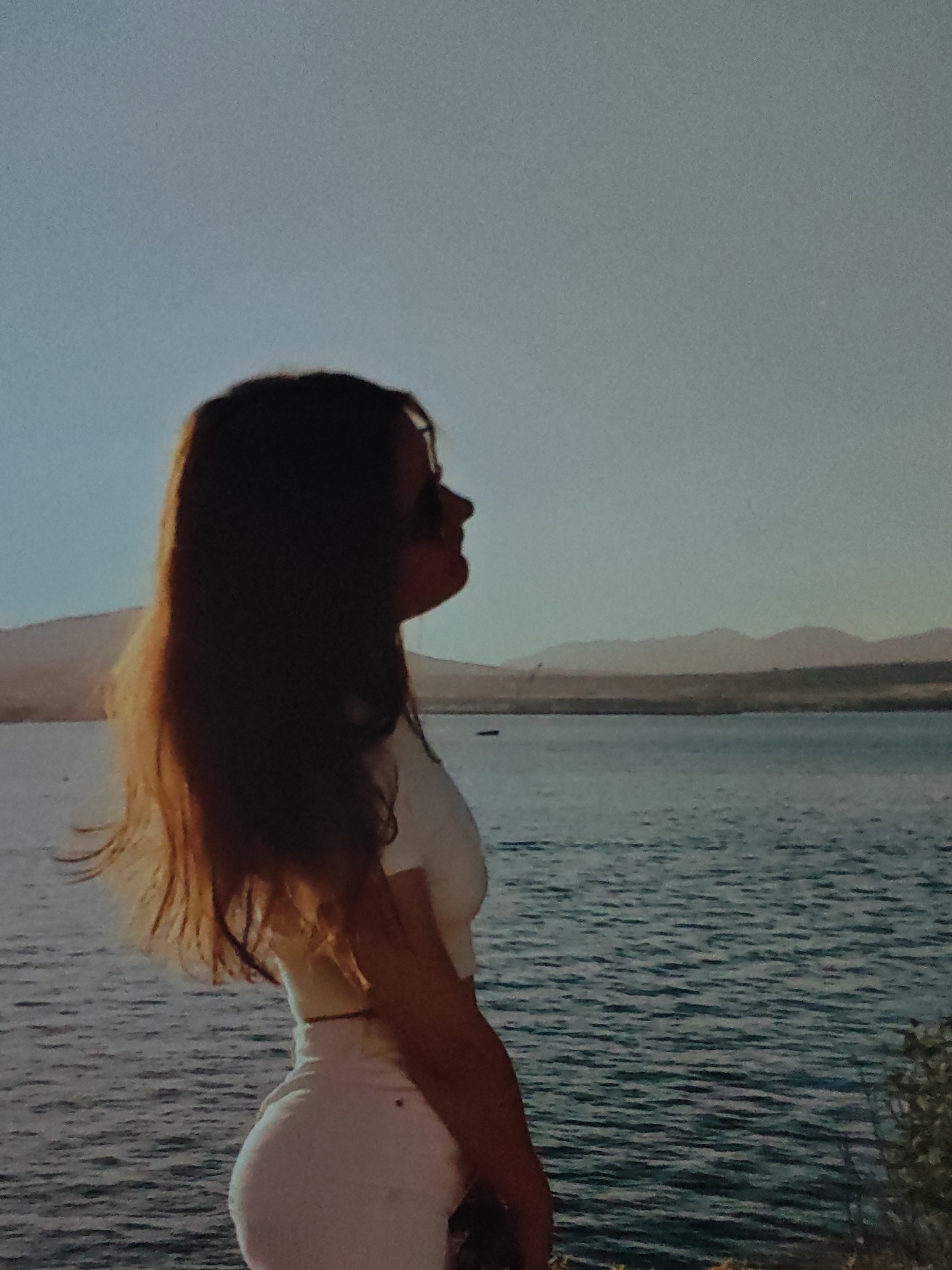 A side profile of a young woman with long, dark hair is captured against a hazy blue sky and a body of water. She is wearing a white crop top and light-colored pants. The background features a distant shoreline with hills or mountains. The lighting suggests it is either early morning or late afternoon.