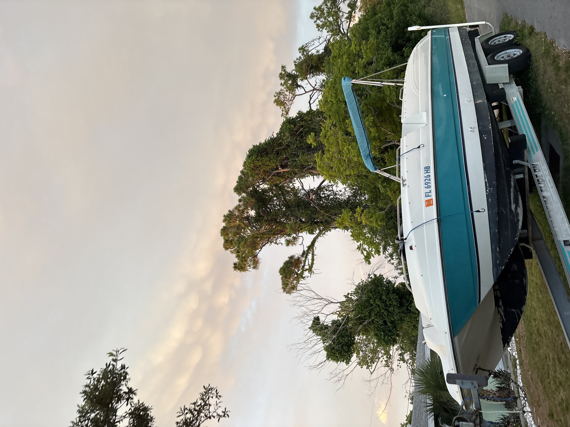 A white and teal speed boat is parked on a trailer on a grassy area next to a paved road. The boat has a blue bimini top and the license plate "FL 6926 HB" is visible on the side of the boat. Behind the boat and trailer are dense green trees and foliage, with a house visible in the background to the left. The sky above is cloudy with hints of pink and orange, suggesting it is either sunrise or sunset. The scene is set in South Venice, Florida, United States of America.