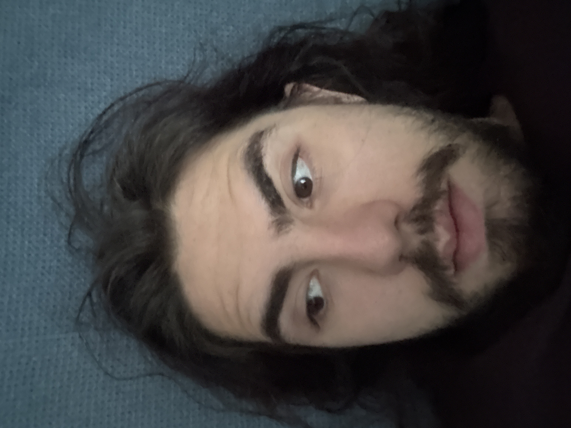 A person with long, dark, messy hair and a beard is lying down, looking to the left. The person's face is illuminated from the right. The background is a textured, teal-colored surface, possibly a blanket or couch. The person is wearing a dark garment, possibly a t-shirt or hoodie.