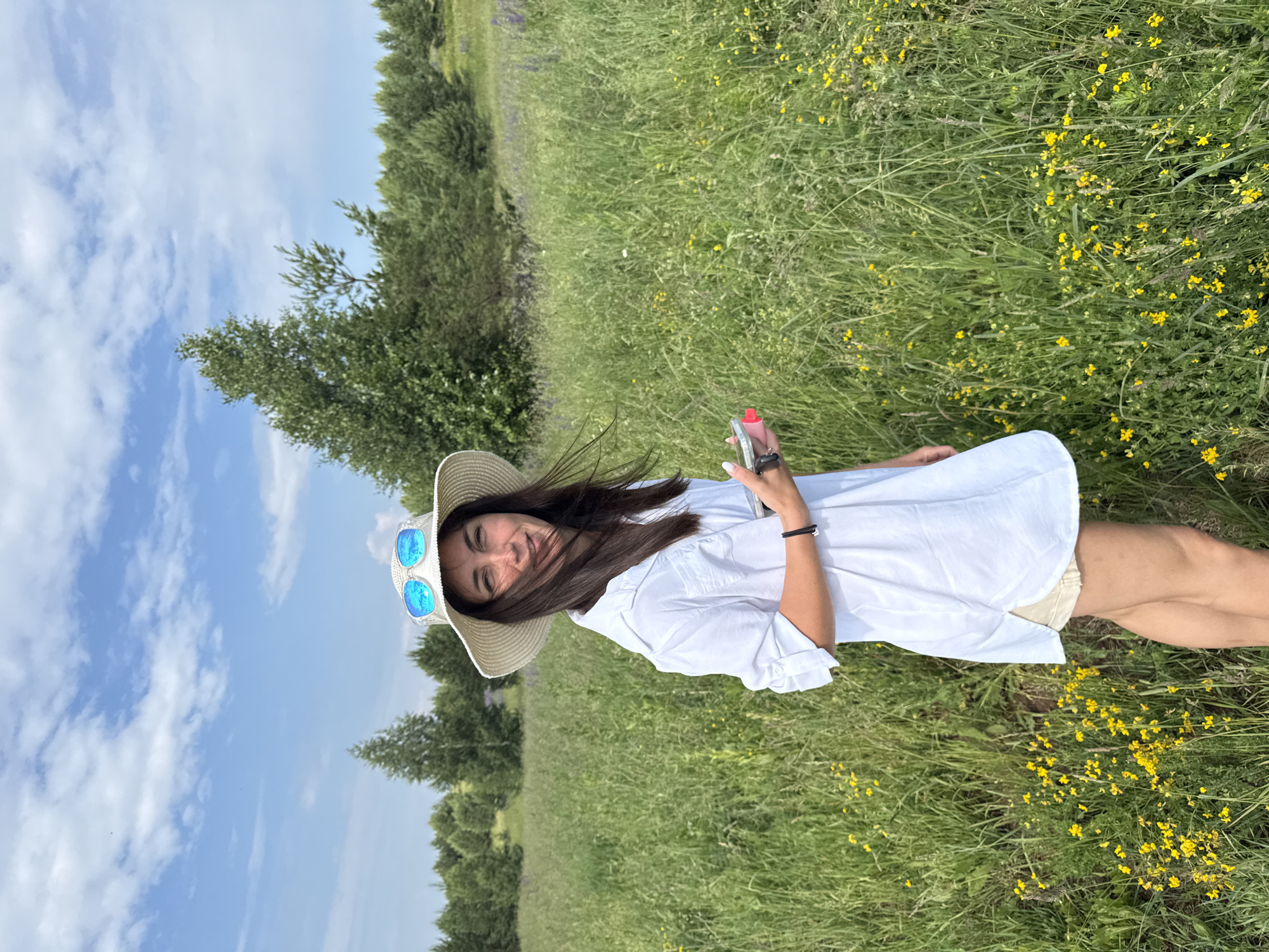 A woman stands in a grassy field wearing a white shirt, shorts, and a straw hat. She is also wearing sunglasses and holding a small, pink object in her right hand. The field is filled with tall grass and small yellow flowers. In the background, there are trees and a blue sky with white clouds. The location is identified as Van'ki, Russia.