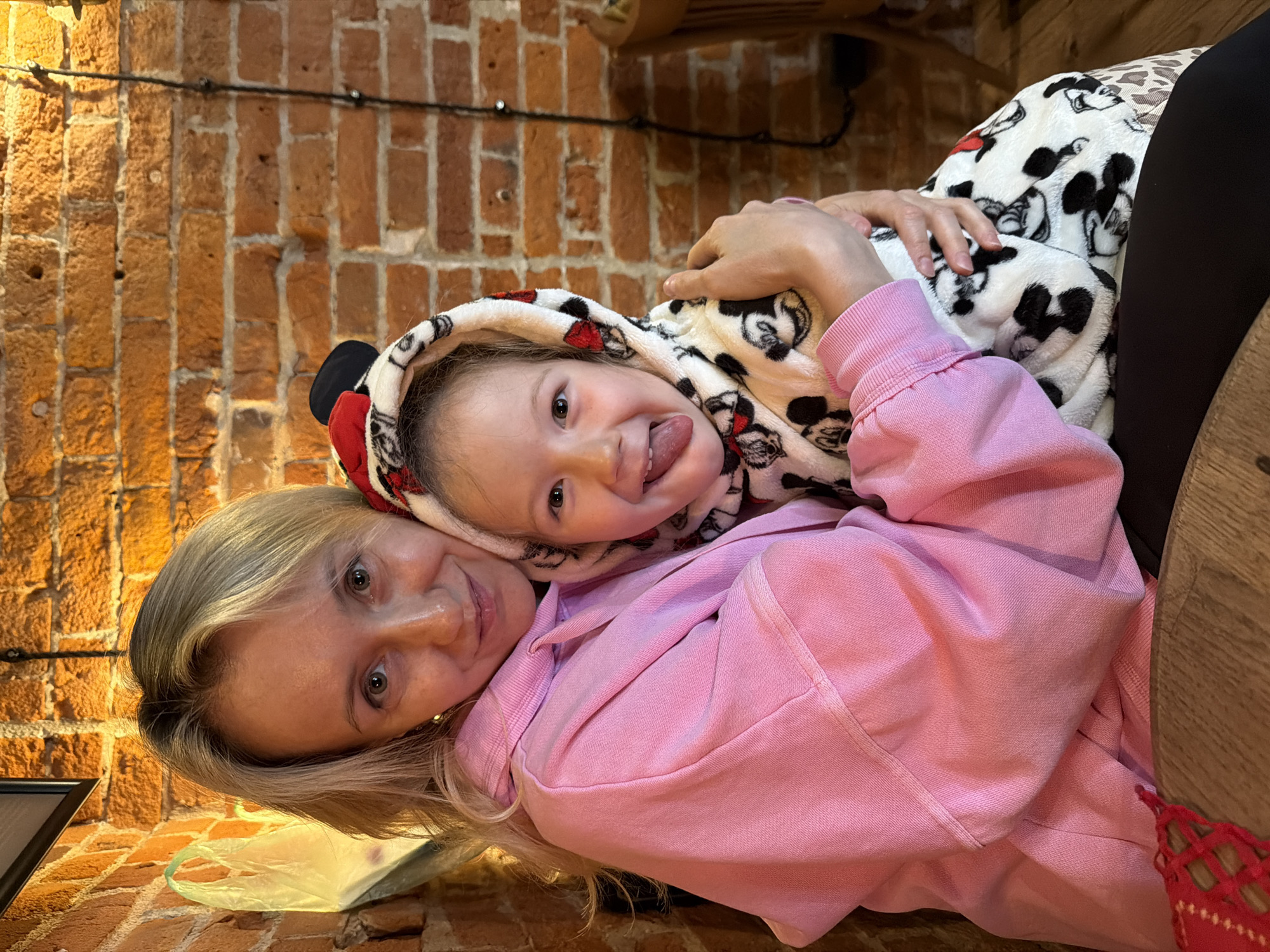 A woman with blonde hair is lying on her side, facing the camera, while a child in a Mickey Mouse hooded blanket lies on top of her, sticking their tongue out. The woman is wearing a pink sweatshirt. The background shows a brick wall. The location is likely indoors. There is no information in the image to indicate it is in Moscow, Russia.