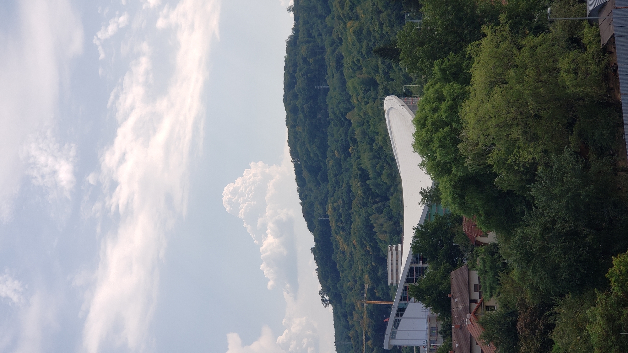 The image captures a daytime scene in Prešov, Slovakia, showcasing a modern stadium under construction nestled against a verdant, tree-covered hillside. In the foreground, a cluster of houses with red tile roofs is partially visible through a dense canopy of trees, suggesting a residential area adjacent to the development. The stadium, characterized by its sweeping white roof and a partially completed facade, dominates the mid-ground, with a construction crane visible to its left. The hillside behind the stadium is densely forested, with power lines subtly integrated into the landscape. The sky above is a mix of blue and white, with several puffy clouds adding depth and texture to the scene. The overall impression is one of urban development encroaching on a natural landscape.