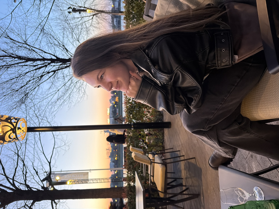 A young adult female is seated in profile, facing right, at an outdoor establishment during sunset or dusk. She has long brown hair and a faint smile, gazing upwards and to the right, with her right hand resting under her chin. She is dressed in a black leather jacket over a dark top, dark grey wide-leg trousers, and dark, lug-soled boots. A brown fanny pack is slung across her front. On a dark table to her immediate left, an empty stemmed glass and a partially visible green glass bottle are present.
The scene is an outdoor paved terrace, featuring additional empty tables and chairs with light-colored cushions behind the subject. A decorative street lamp with an illuminated, ornate metal fixture stands in the mid-ground. Bare deciduous trees are visible against the sky. In the background, beyond a low hedge, a body of water is visible, with the illuminated skyline of buildings on the opposite bank. A vertical banner in the mid-ground displays text including "BELGRADE WATERFRONT" and "DIREKCIJA", identifying the location as Belgrade Waterfront along the Sava River in Belgrade, Serbia. Several indistinct figures are visible near the waterfront in the distance. The sky exhibits a gradient from orange at the horizon to light blue overhead.