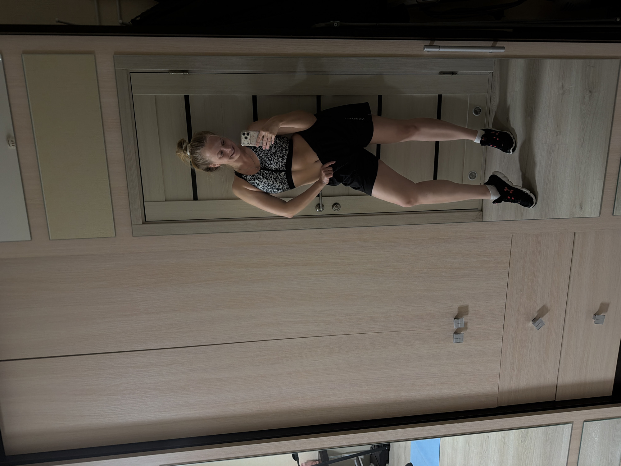 A young adult female is captured in a full-length mirror selfie, presumably taken indoors in the city of Izhevsk, Russia. She stands smiling, holding a light-colored smartphone in her right hand to take the photo. Her left hand is hooked into the waistband of her black athletic shorts, slightly pulling them down. She is wearing a black and white patterned sports bra, white athletic socks, and black sneakers with red accents. Her blonde hair is tied back in a bun. The background visible in the mirror includes a light-colored wooden wardrobe or closet with silver square handles on drawers and doors, and a light wooden interior door with a silver handle. The flooring is light wood. A reflection of a blue mat and dark furniture is visible on the far left side of the mirror. A small white circular sensor or detector is visible reflected near the top of the wall above the door.