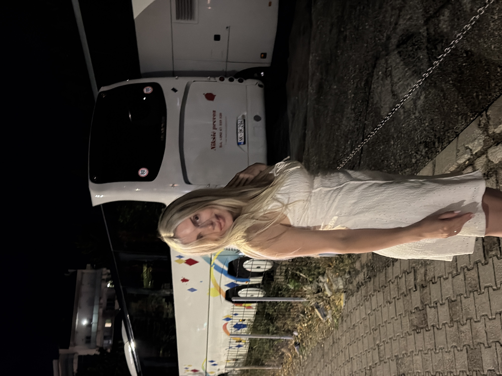 A young woman with blonde hair is standing next to a white bus. She is wearing a white dress and has her left hand on her hip. The bus has a logo with a red apple and text that reads "Niksic prevoz" and "Tel. +420 415 939 528". The license plate on the bus is "MK-BG 252".
The scene appears to be outdoors at night, possibly a parking lot or roadside, given the presence of a paved walkway and a gravelly area. A metal chain is visible in the background, and some foliage is present. The woman's expression is a slight smile, suggesting a casual or pleasant moment. The bus also has colorful diamond-shaped designs on its side. The location is specified as Budva, Montenegro.