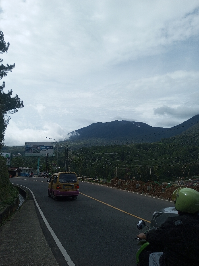 The image depicts a mountainous road scene in the city of Sandamal Eliya, Sri Lanka, under an overcast sky. In the foreground, the back of a person wearing a bright lime-green full-face helmet and a dark jacket is visible, operating a green scooter or small motorcycle. The vehicle is positioned on the right side of a two-lane asphalt road, which features a solid white line on the shoulder and a broken yellow center line, curving slightly to the left. Further down the road, a yellow and purple minivan drives away from the viewer.
To the left of the road, a steep, green, vegetated embankment with a concrete drainage ditch runs parallel to the asphalt. A tall conifer tree is partially visible at the far left edge. Adjacent to the road on the left, a large billboard with light blue supports displays an advertisement, partially reading "DPRK CLOUD". A street light pole stands near the billboard. Small, simple buildings and parked motorcycles are visible in the distance beyond the billboard, suggesting a roadside cluster.
The right side of the road shows a sloped area with disturbed brown earth and small, newly planted saplings, some with white protective wrapping around their bases. Multiple utility wires or power lines traverse the midground horizontally. In the background, dark green, densely forested mountains rise under the cloudy sky, with some peaks partially obscured by low-hanging mist or clouds. Distant residential structures are faintly visible at the base of the mountains on the right.