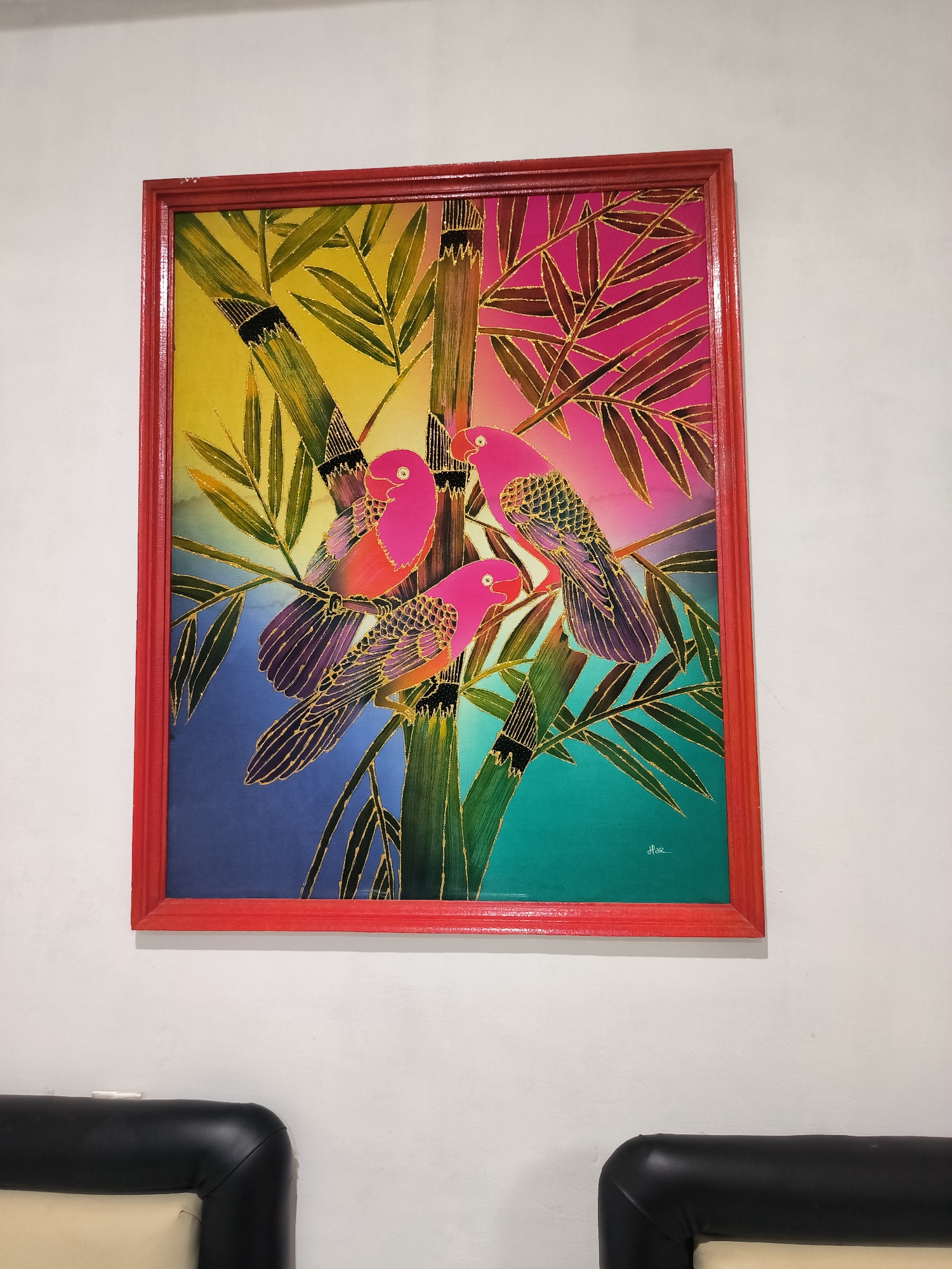 The media file displays a vibrant framed painting hanging on a plain white wall, with parts of two cushioned chairs visible below it. The painting, likely a batik artwork given its style, features three brightly colored pink birds with patterned wings, perched among bamboo stalks and leaves.
The background of the painting is a striking gradient, transitioning from yellow in the upper left to pink in the upper right, then blending into blue in the lower left and teal in the lower right. All elements within the painting – the birds, bamboo stalks, and leaves – are distinctly outlined in gold, which adds a decorative and shimmering effect. The birds themselves are predominantly pink, with details on their wings showing shades of purple and black. The bamboo stalks are green with black bands, and the leaves are also green, interspersed with the gold outlining.
The painting is encased in a prominent, textured red wooden frame. In the bottom right corner of the artwork, a stylized signature "Hoo" is visible. Below the painting, in the foreground, are the rounded corners of two pieces of furniture, likely chairs or sofas, upholstered in black and cream or white material. The overall setting appears to be indoors, possibly a living room or lounge area in Banda Aceh, Indonesia, suggesting the cultural context of the artwork. No specific activity is taking place beyond the display of the art.
