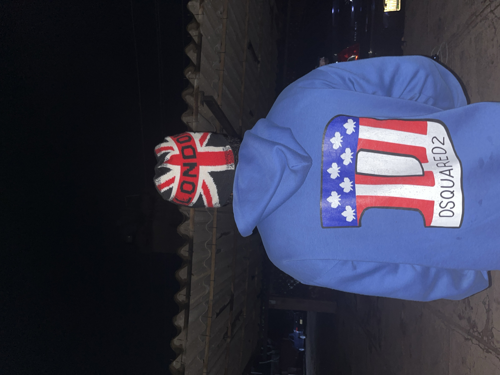 The image shows the back of a person wearing a blue hoodie with a large graphic design on it and a beanie that resembles the Union Jack flag, with "LONDON" written on it. The graphic on the hoodie features red and white stripes with blue accents and white leaf-like shapes, and the text "DSQUARED2" below it. The person is standing outdoors at night, with a corrugated metal roof structure visible behind them. In the background, a dark-colored car is parked. The scene appears to be in a dimly lit, possibly outdoor residential or commercial area. There is no specific visual information in the image that indicates the location is in Jos, Nigeria.