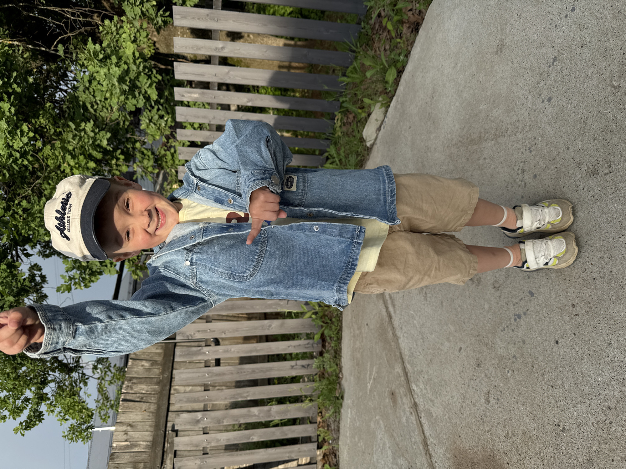 A young boy, approximately 6 years old, stands on a concrete pathway in front of a wooden fence. He is wearing a light blue denim jacket over a yellow t-shirt, tan shorts, a beige baseball cap with dark blue accents, and white sneakers with yellow and grey detailing. The boy is smiling and has his left arm raised with his fist clenched. The background features green trees and foliage, and the lighting suggests it is daytime with sunlight filtering through the leaves. The setting appears to be a residential area, possibly a backyard or a street in Chaikovsky, Russia.