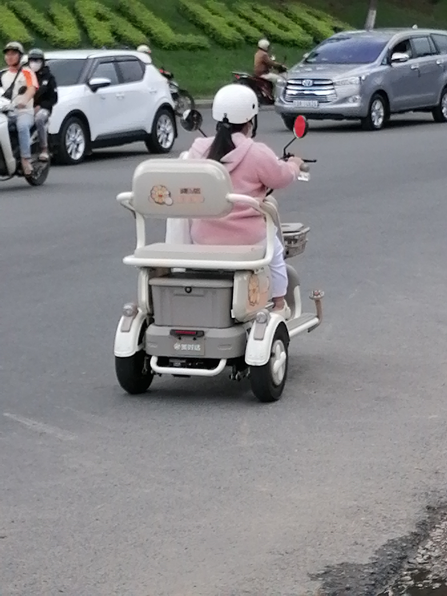 The image captures a street scene in Da Lat, Vietnam, featuring several vehicles and pedestrians. In the foreground, a person on a three-wheeled electric scooter, wearing a pink hoodie and a white helmet, is moving away from the camera. The scooter has a grey storage box and decorative stickers. To the left of the scooter, a white car is parked, and two people are on a motorcycle, one driving and the other a passenger wearing a white helmet and mask. In the background, a silver car is driving on the road, and another person on a motorbike is also visible. The environment appears to be a paved road with green foliage on a slope in the upper part of the image, suggesting an urban setting within Da Lat.