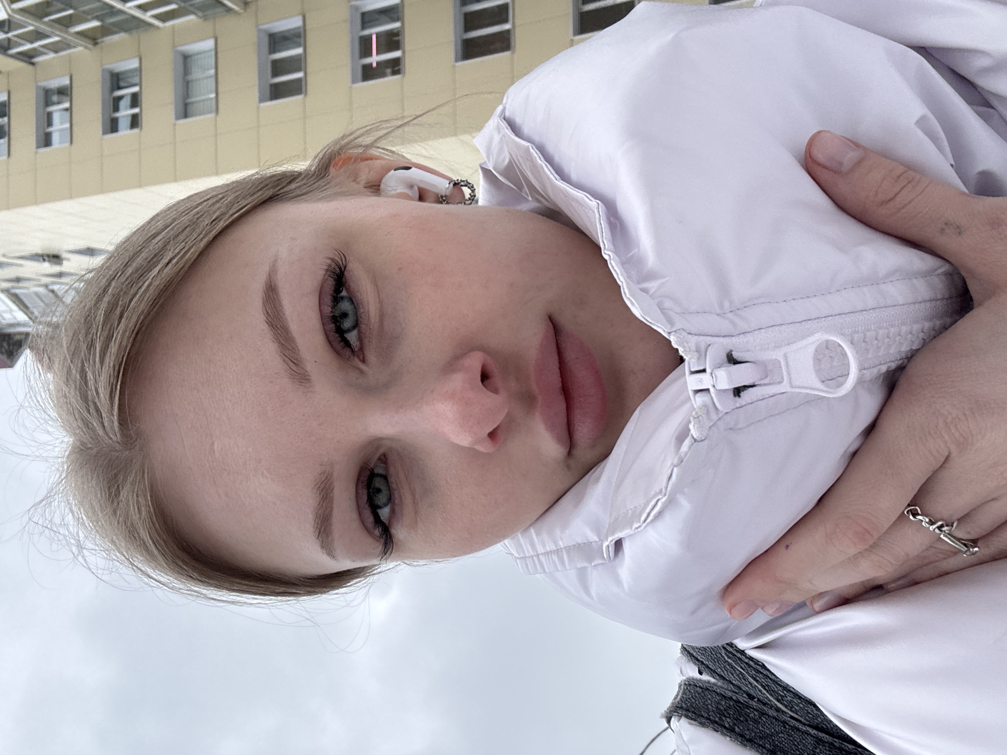 A young woman is taking a selfie outdoors in Perm, Russia. The photograph is taken from a slightly downward angle, focusing on her face and upper body. She has light blonde hair, fair skin, and green eyes, and is wearing a white puffer jacket. She has an earbud in her left ear, and a silver ring on her right hand. The background features a multi-story building with rectangular windows, typical of urban architecture. The sky is overcast, suggesting a cloudy day in the city.