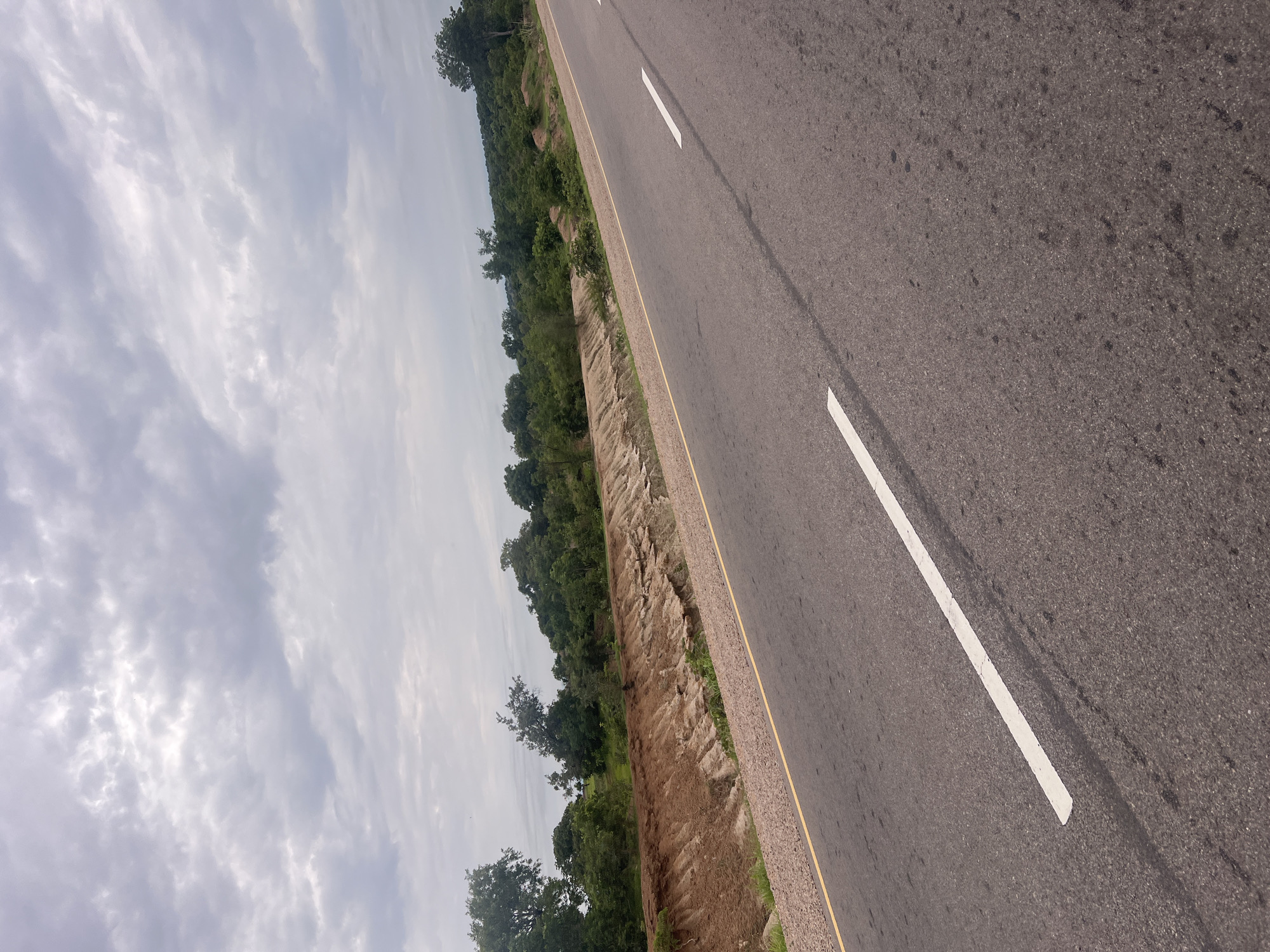 This media file is a landscape photograph taken from a low angle, looking down a highway. The road surface is asphalt, marked with a solid white line on the right side. A yellow line runs along the edge of the road. To the left of the highway, there is a steep embankment covered in dry earth and vegetation. Beyond the embankment, a dense forest of green trees stretches into the distance, with some larger trees visible in the foreground. The sky is overcast with white and gray clouds, suggesting it is daytime. The overall scene depicts a rural highway in Minda, Nigeria, possibly after recent rainfall given the appearance of the embankment. There are no people or text visible in the image.