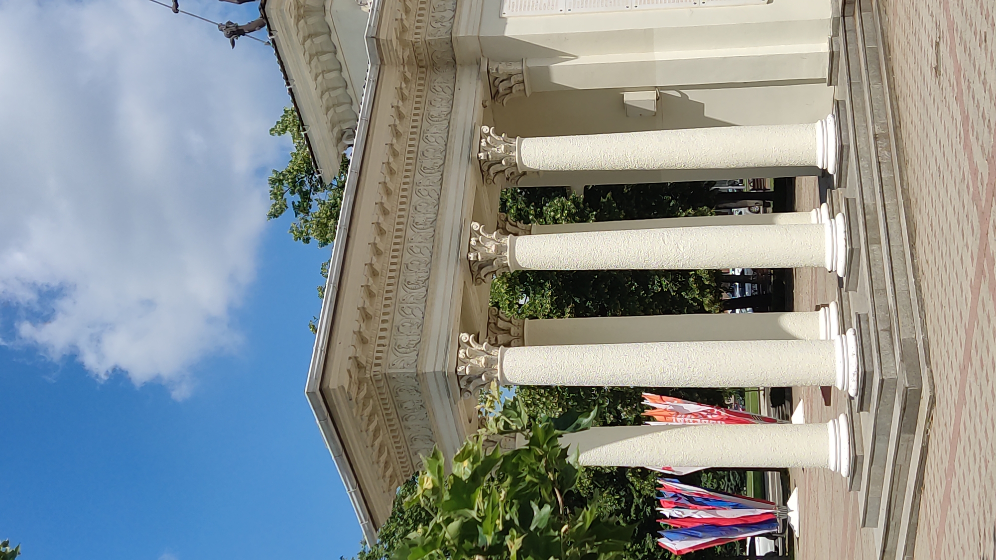This is a vertical medium shot of a white colonnade with intricate carvings above the columns. The colonnade is part of a building's facade, which is light beige in color. The columns are fluted and topped with ornate capitals. Behind the columns, lush green trees are visible. In the foreground, a cluster of small, colorful flags are strung together. These flags appear to be red, white, and blue. The sky above is a clear, bright blue with a few scattered white clouds. The scene suggests a sunny day, likely late morning or early afternoon given the shadows cast by the colonnade. The paving stones leading up to the building are visible in the lower right portion of the image. There is no visible text in this image. The setting appears to be an outdoor public space, possibly a park, museum, or government building, in Krasnodar, Russia. The overall impression is one of architectural grandeur and a festive atmosphere due to the flags.