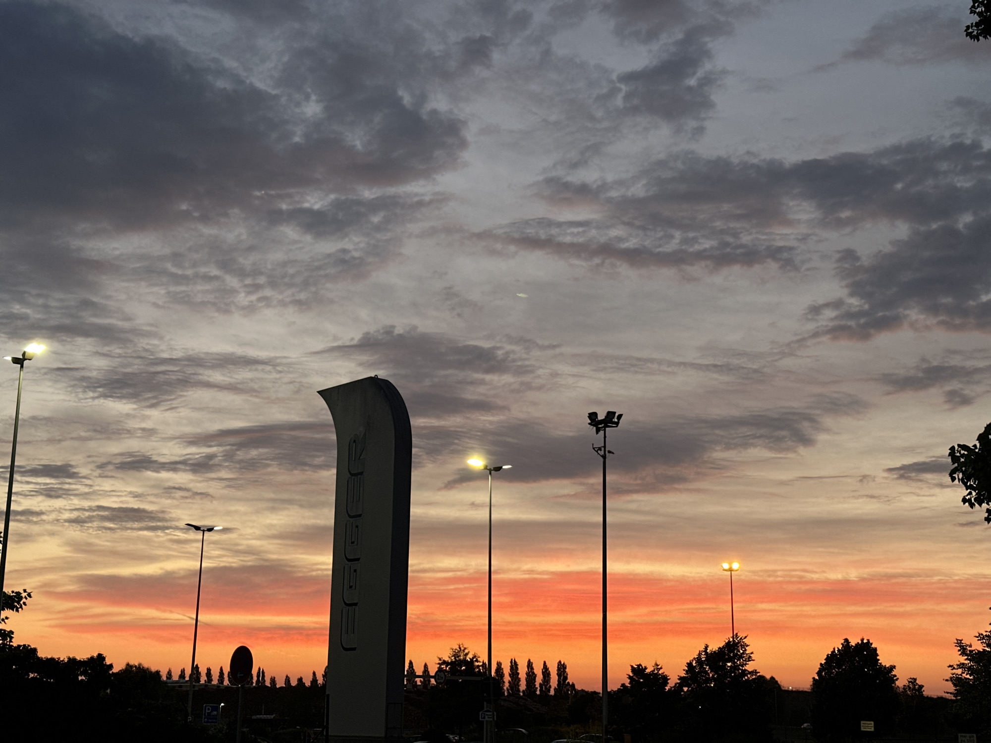 This is an image of a sunset in Wismar, Germany. The sky is filled with clouds in shades of gray, pink, and orange, with the orange hues concentrated near the horizon. In the foreground, there is a tall, slender sign with the word "EGEER" written vertically in white. Several lampposts are visible, some illuminated, adding to the scene. Silhouetted trees and a few indistinct structures form the dark landscape beneath the vibrant sky. The overall impression is of an outdoor scene, possibly a parking lot or a commercial area, at dusk.