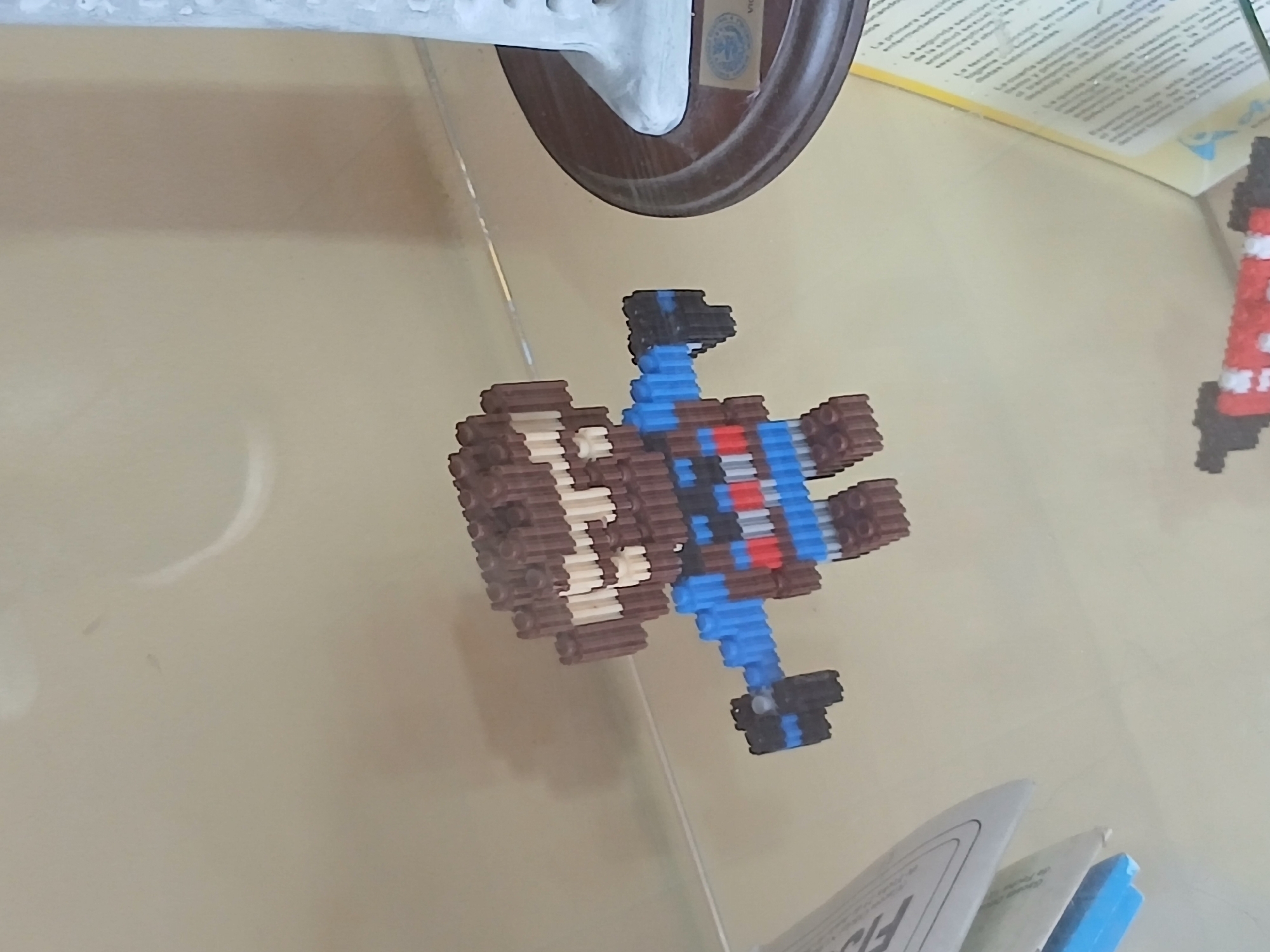 In Juan Griego, Venezuela, a small, brown, pixelated figurine is displayed on a glass surface. The figurine, constructed from interlocking blocks, resembles a character with a large mouth and what appear to be white teeth. It has blue arms with black ends and a body detailed with blue and red stripes. To the right of the figurine, a portion of a white, lighthouse-shaped object on a dark circular base is visible. The background is a neutral, light-colored surface.