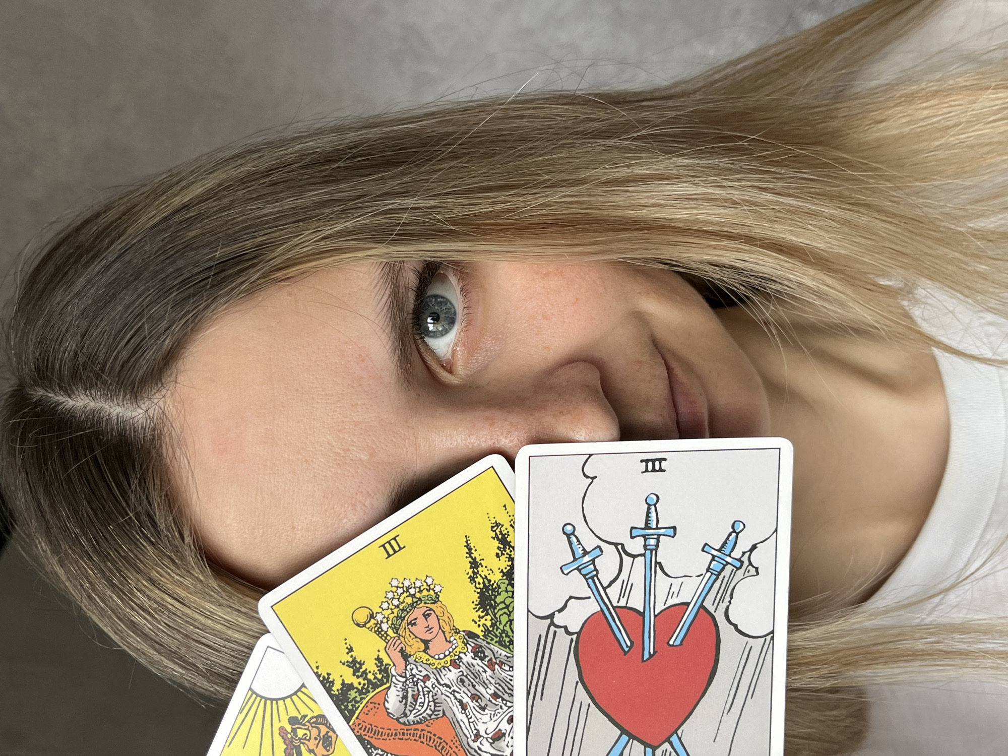 A woman with blonde hair and blue eyes is holding up tarot cards to her face. One card is the Three of Swords, depicting a heart pierced by three swords. Another card is the Queen of Wands. The image's location within the city of Chaikovsky, Russia, is not discernible from the provided content.