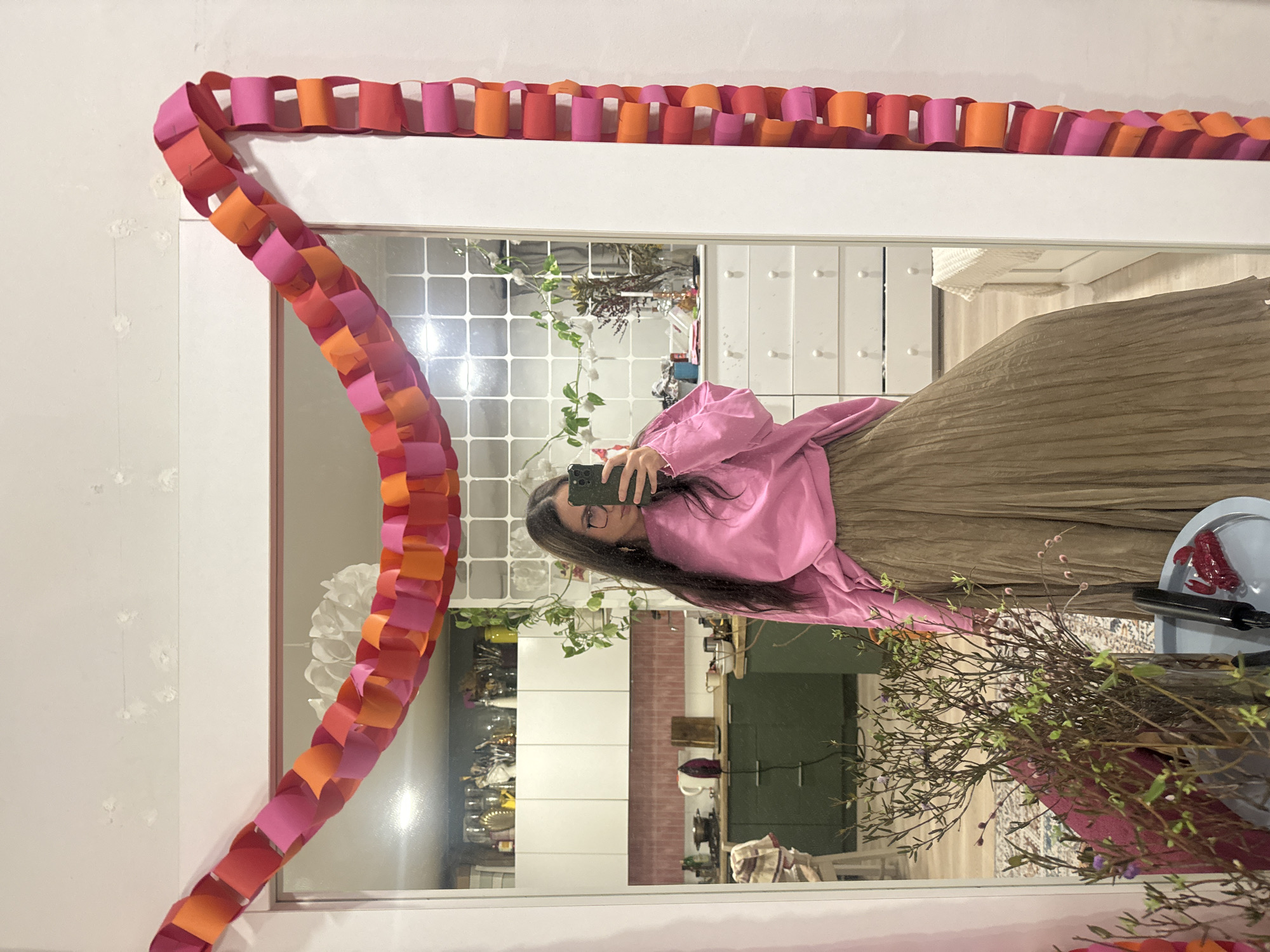 A woman captures her reflection in a mirror selfie within an indoor setting, likely a home in Krasnoyarsk, Russia. The image is rotated 90 degrees clockwise. She has long, dark hair, wears glasses, a bright pink long-sleeved top, and a pleated olive green or brown skirt. She holds a dark-colored smartphone to take the picture.
The large, rectangular mirror has a white frame, which is decorated along its top and left side with a colorful paper chain garland featuring alternating links of pink, orange, and red. The mirror's reflection reveals an interior space, likely a kitchen or living area, with both white and dark green cabinets, a section of exposed brick wall, and various household items such as a kettle, jars, and cups. Decorative elements include several potted plants or branches with small leaves and buds, and a large, light-colored ceramic or paper flower. A light blue bowl holding a red lobster figurine and a black marker pen is also visible in the reflection. A white grid pattern appears on what may be a window or door in the reflected background. The wall to the immediate left of the mirror is white and shows some minor imperfections. No discernible text is visible anywhere in the image.